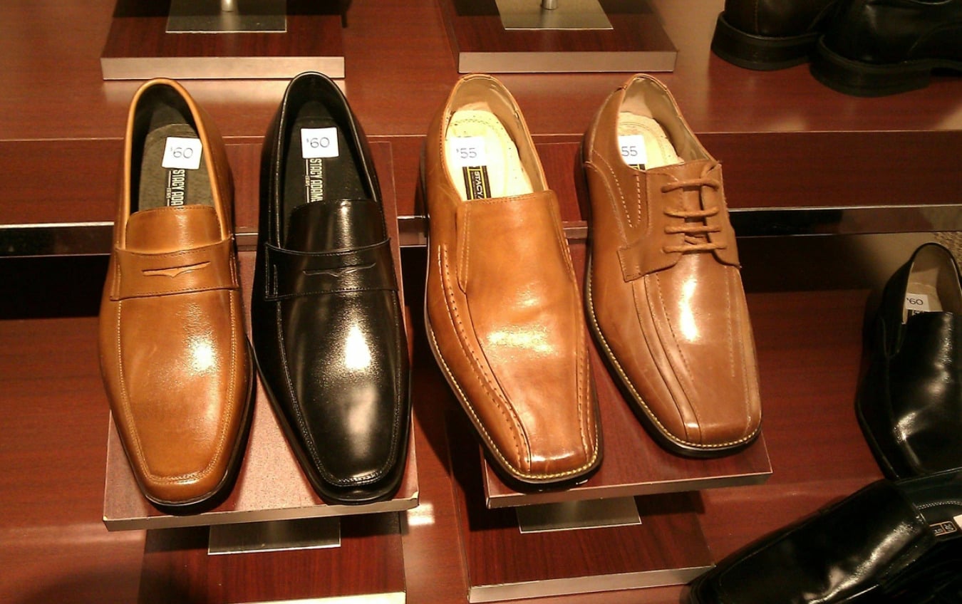 Three pairs of men's dress shoes, two brown and one black, are displayed on a wooden shelf. The shoes are tagged with prices and neatly arranged in a shoe store.