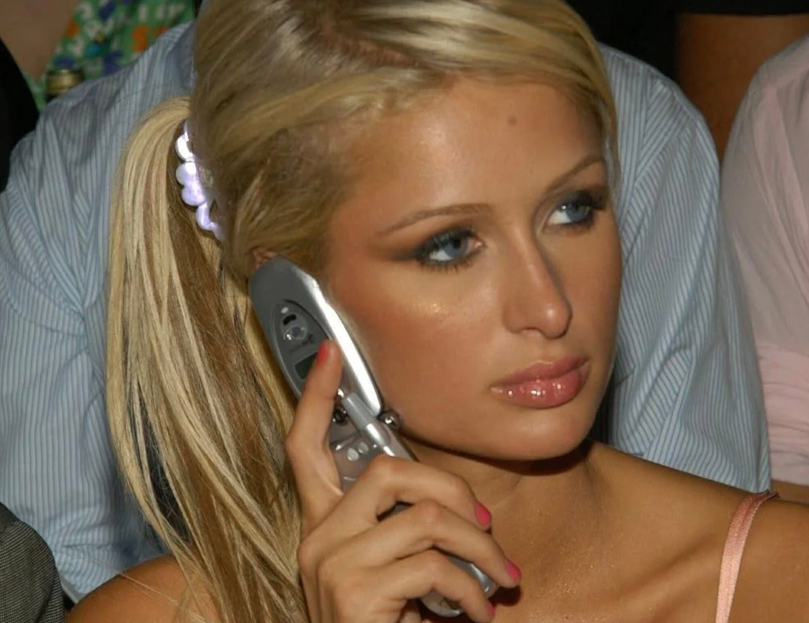 A woman with long blonde hair in a ponytail holds a silver flip phone to her ear. She has light makeup, glossy lips, and wears a pink top. The background is out of focus, with people nearby.