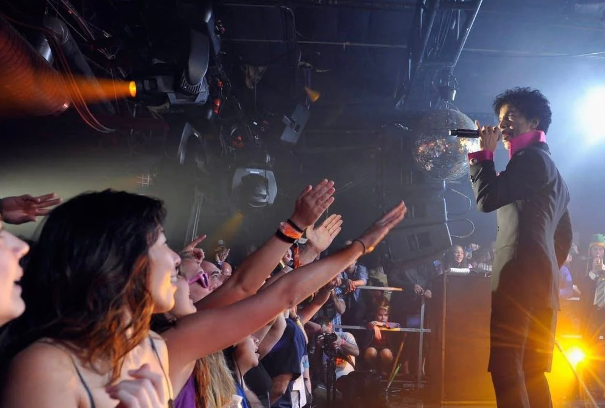 A singer in a black suit with a pink shirt performs on stage, holding a microphone. Audience members reach toward the stage, smiling and cheering under dramatic stage lighting.