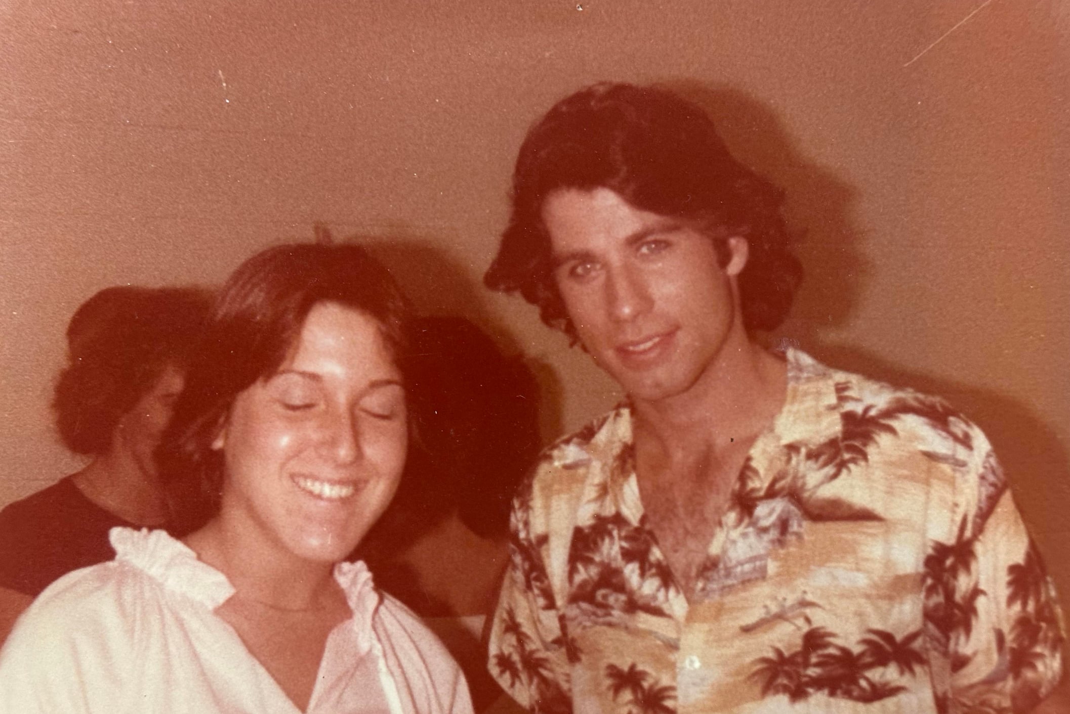 A woman with short brown hair and a white blouse stands smiling with her eyes closed next to a man with wavy dark hair in a tropical print shirt; both are indoors against a plain, light-colored wall.