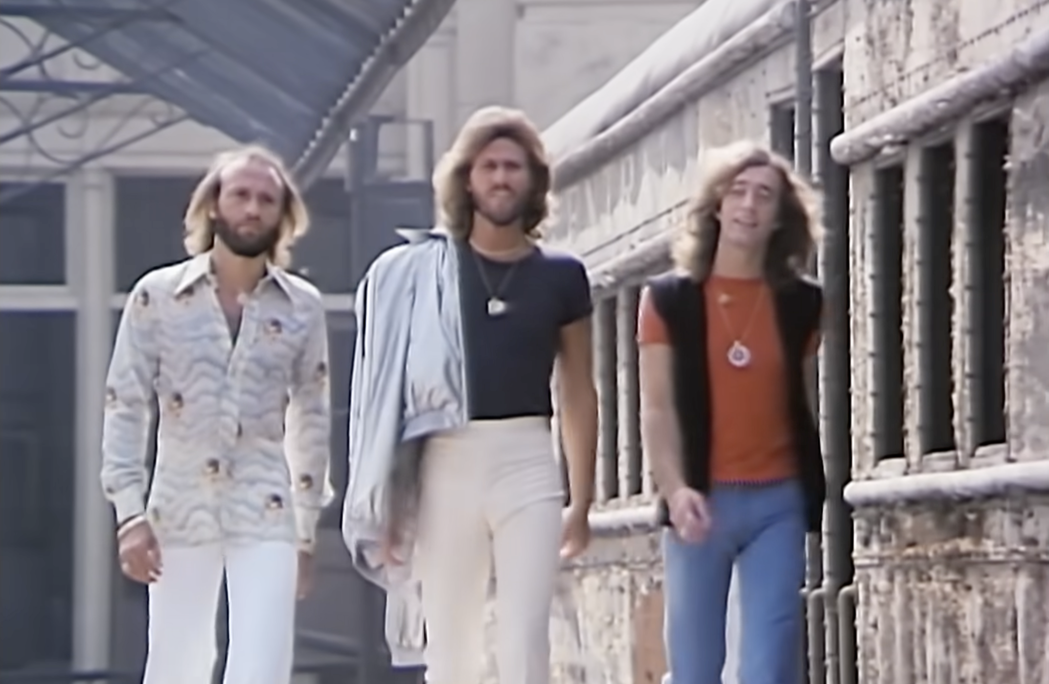 Three men with long hair walk confidently beside a vintage train; one wears a patterned shirt, one a dark shirt with a jacket over his shoulder, and one an orange shirt with a black vest. The setting appears to be an old railway platform.