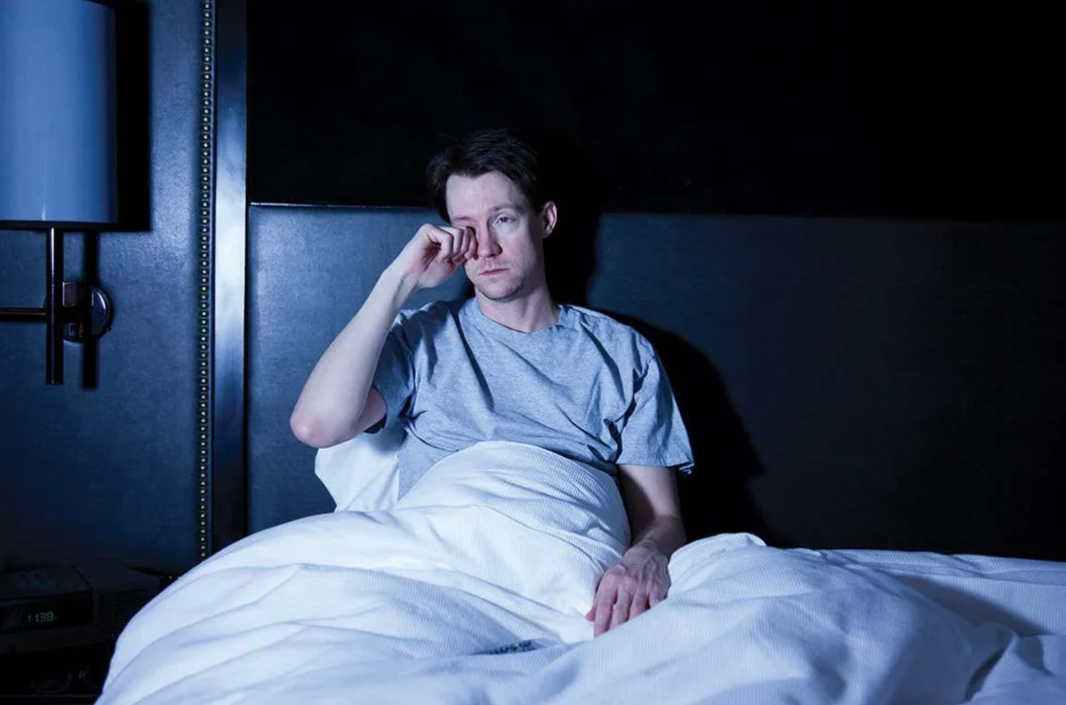 A man sits up in bed against a dark headboard, wearing a gray T-shirt and covered with a white blanket. He looks tired and is rubbing one of his eyes with his hand. The room is dimly lit.