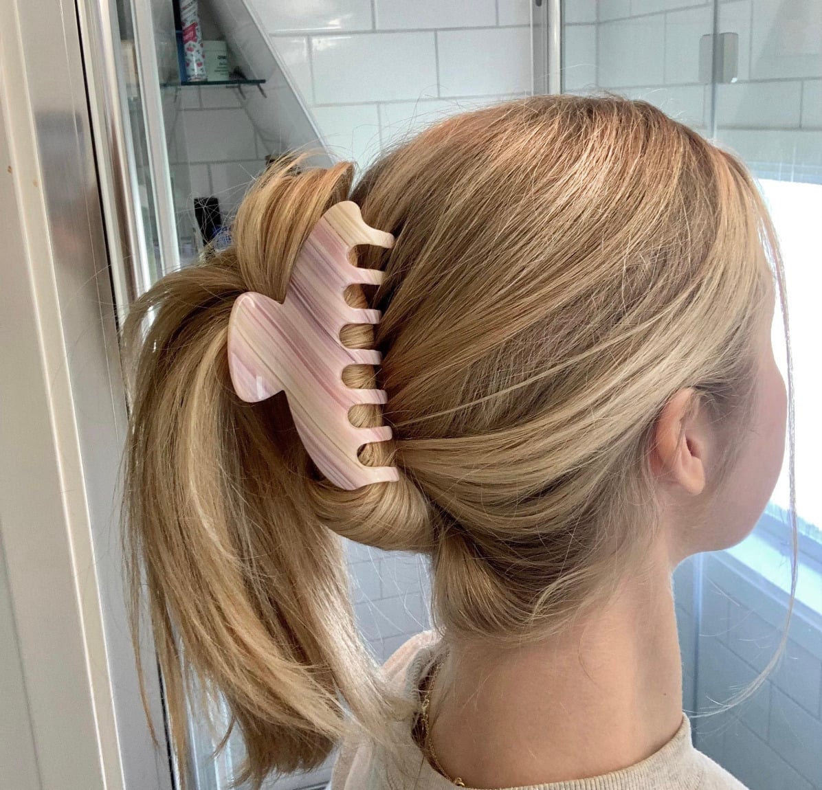 A woman with blonde hair styled in a low twist secured by a large, light pink hair claw clip, standing in a bathroom with white tiled walls.