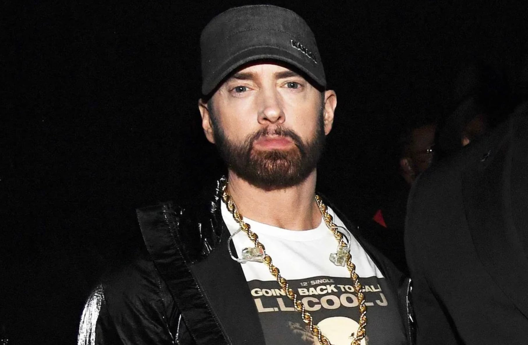 A man with a dark beard wears a black cap, a gold chain, a white T-shirt with a graphic print, and a shiny black jacket, standing against a dark background.