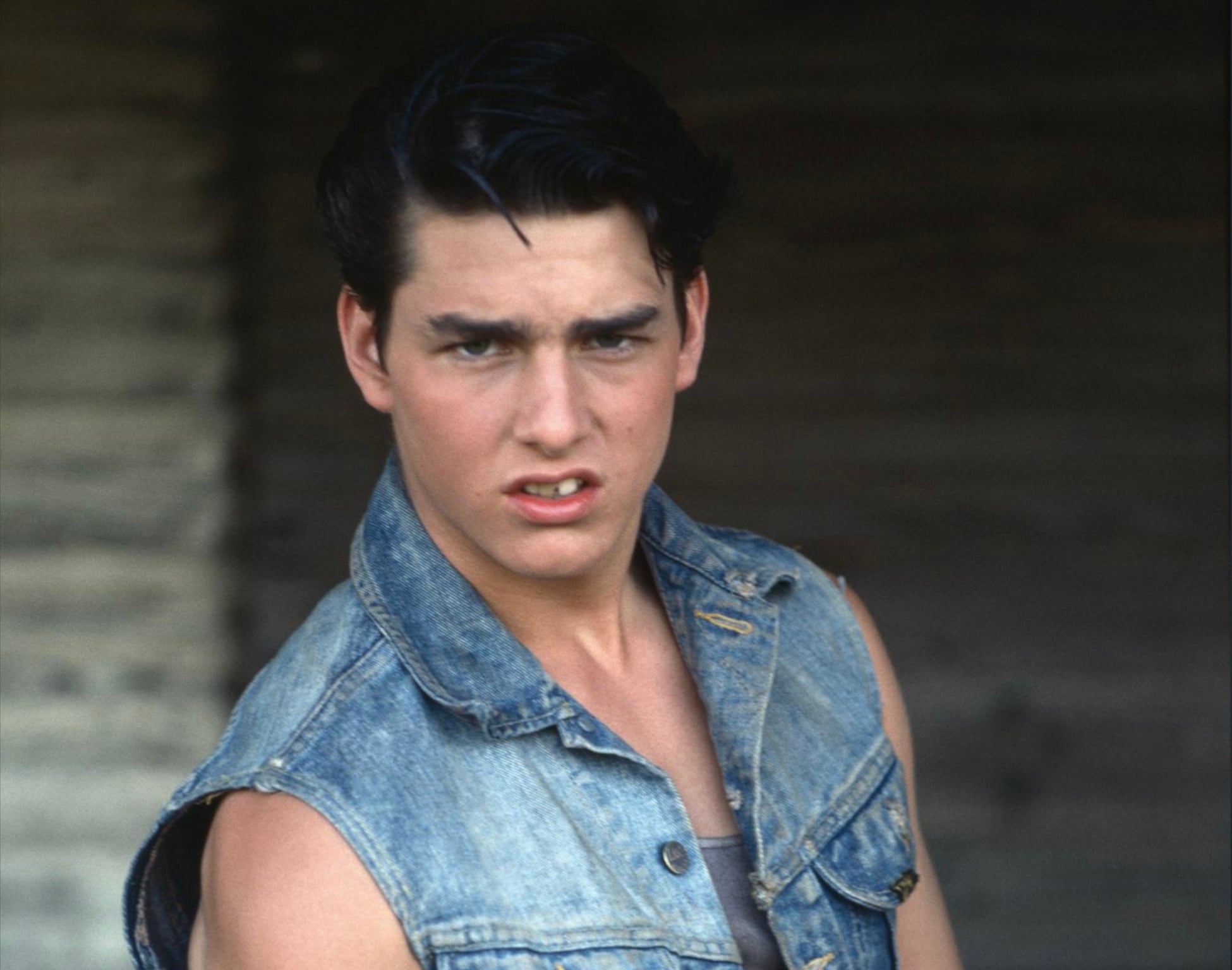 A young man with dark hair wears a sleeveless denim jacket and looks intently at the camera. The background is blurred and wooden, creating a dramatic effect.