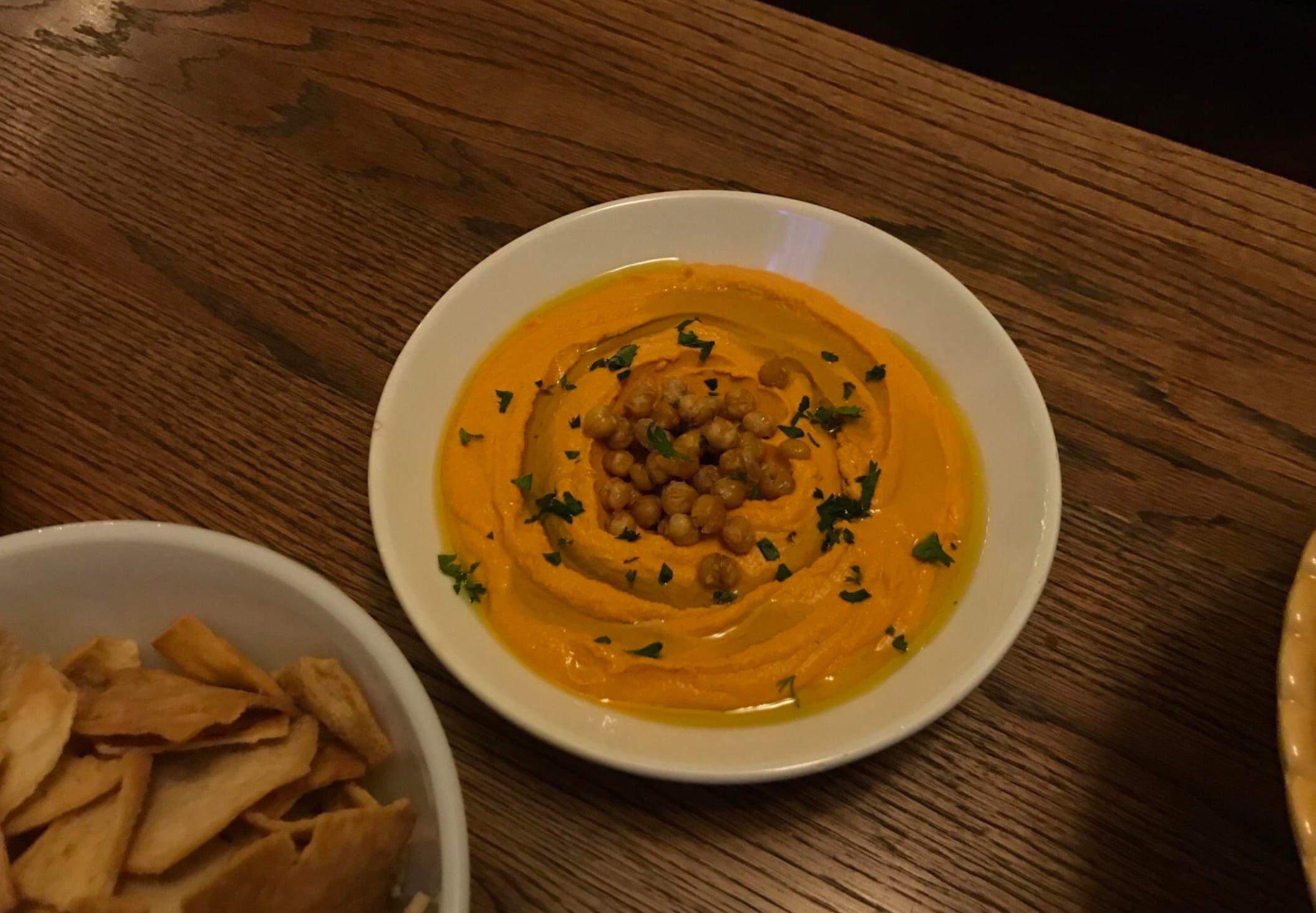 A white plate filled with creamy orange hummus, topped with roasted chickpeas and chopped herbs, sits on a wooden table next to a bowl of pita chips.
