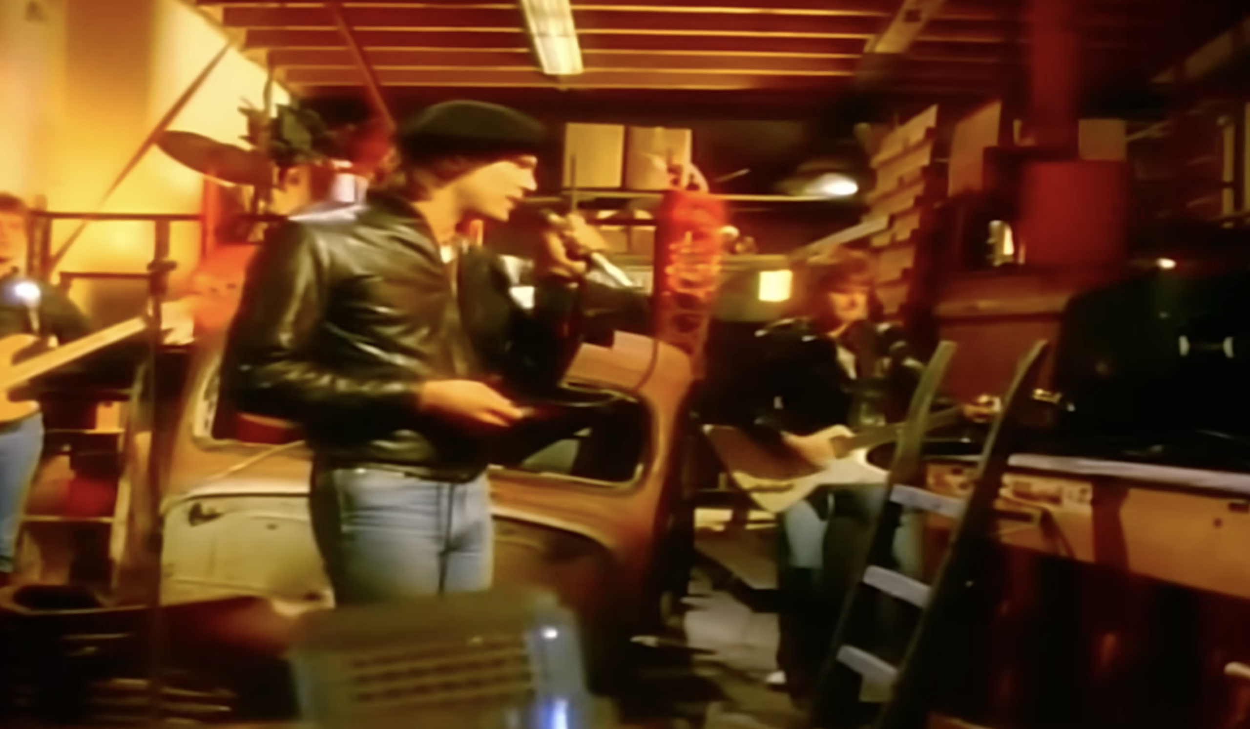 A band performs in a cluttered garage filled with car parts and shelves; the lead singer holds a microphone and wears a black leather jacket and cap, while other members play guitars in the background.