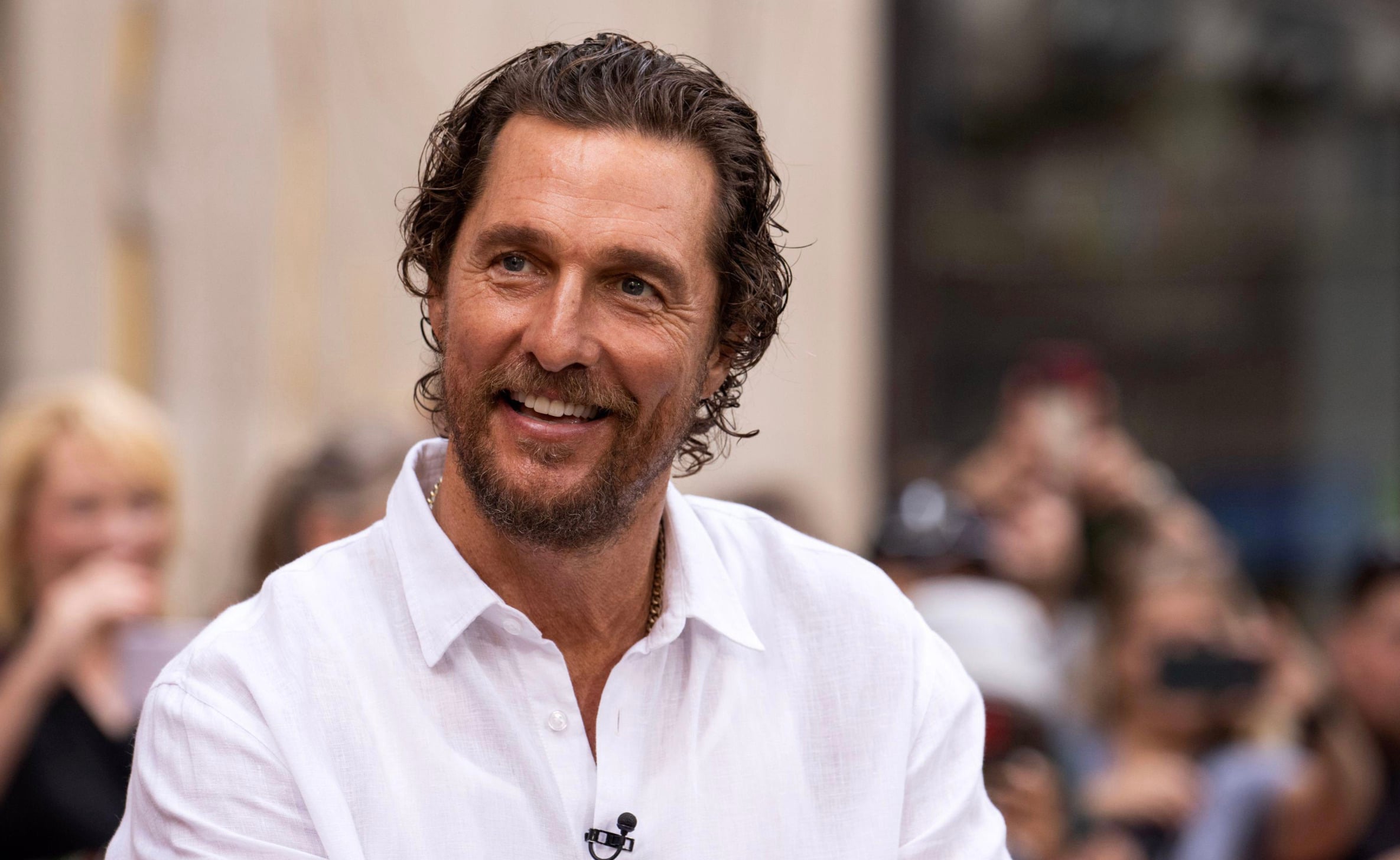 A man with wavy brown hair and a beard smiles while wearing a white button-up shirt outdoors, with a blurred crowd in the background.