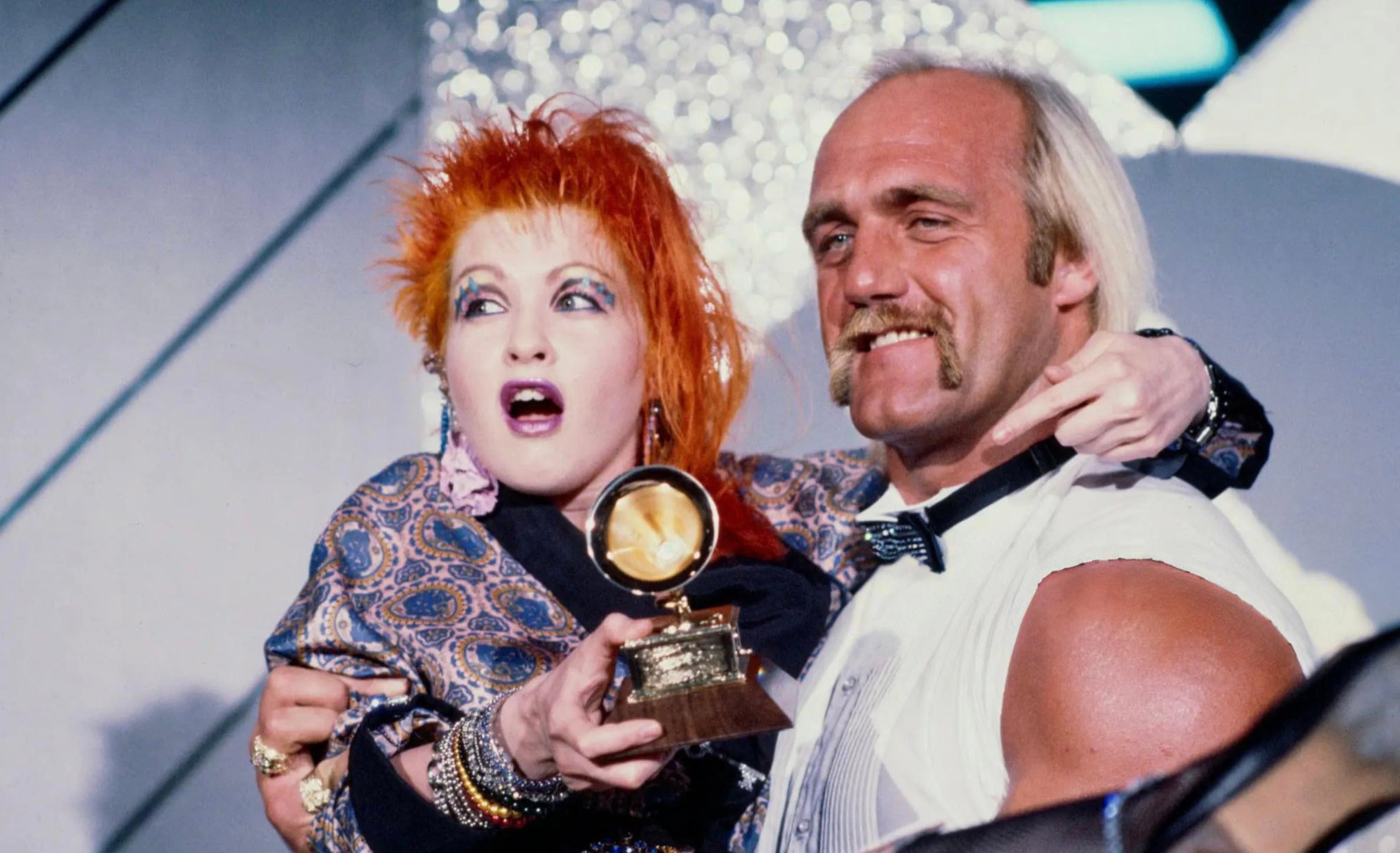 A woman with bright orange hair holds a Grammy award while being hugged by a muscular, mustached man in a sleeveless shirt and bow tie. Both are smiling on stage with a sparkly background.