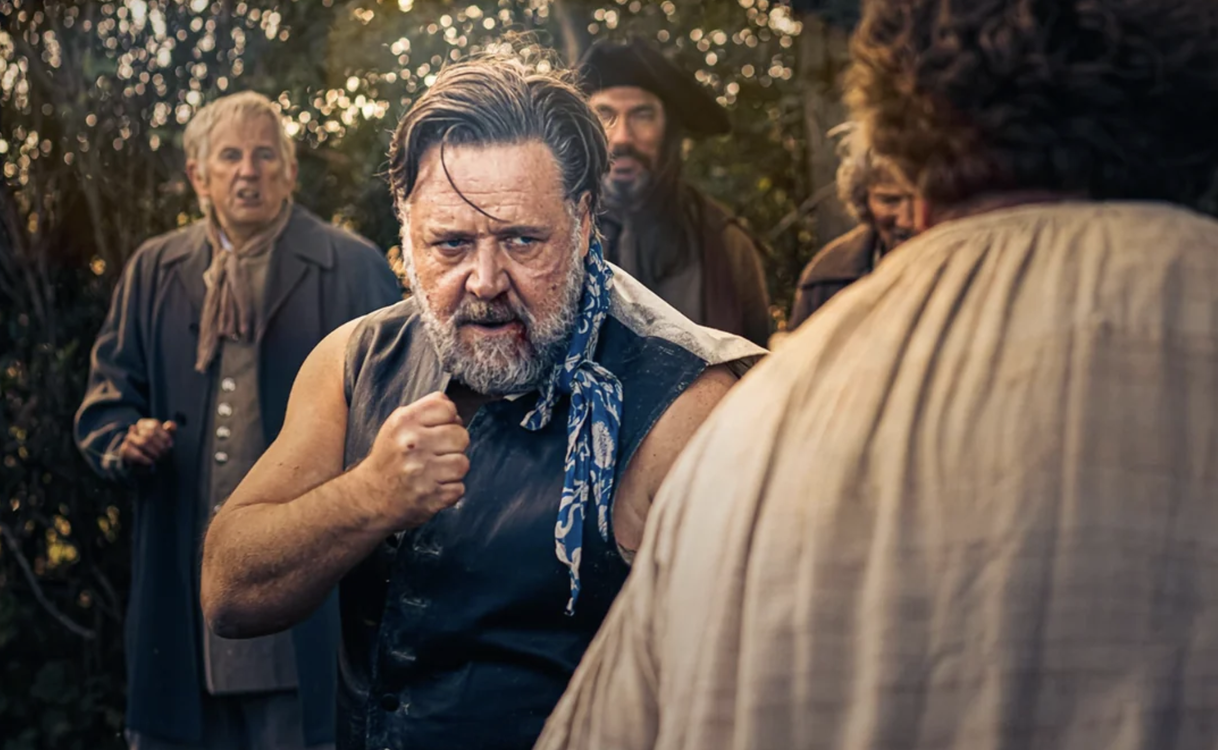 A bearded man in a leather vest with a blue patterned scarf around his neck stands with a clenched fist, facing another person, outdoors, while several people watch in the background.