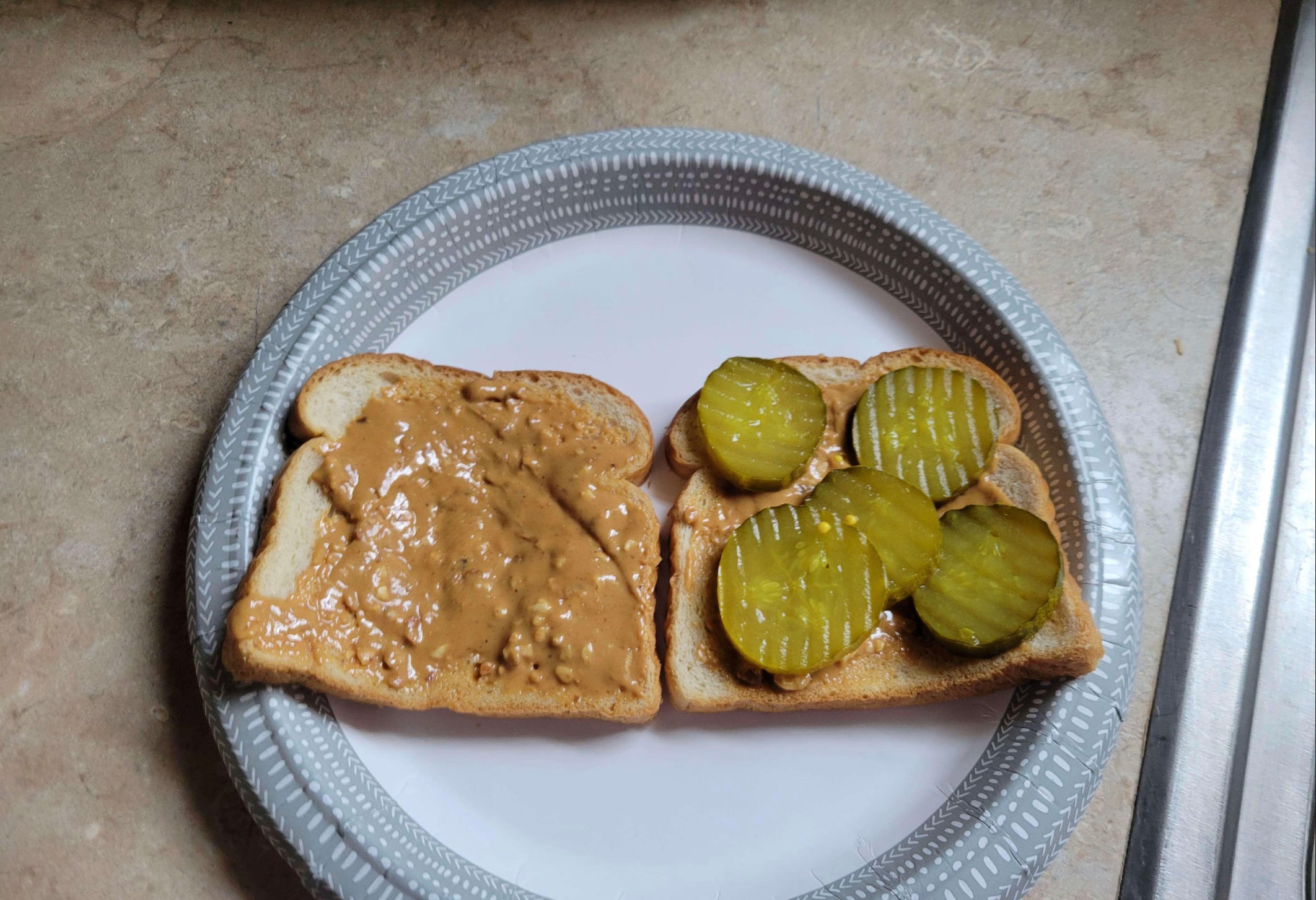 Two slices of toasted bread on a white paper plate; one slice has chunky peanut butter, and the other is topped with several pickle chips. The plate sits on a beige countertop.