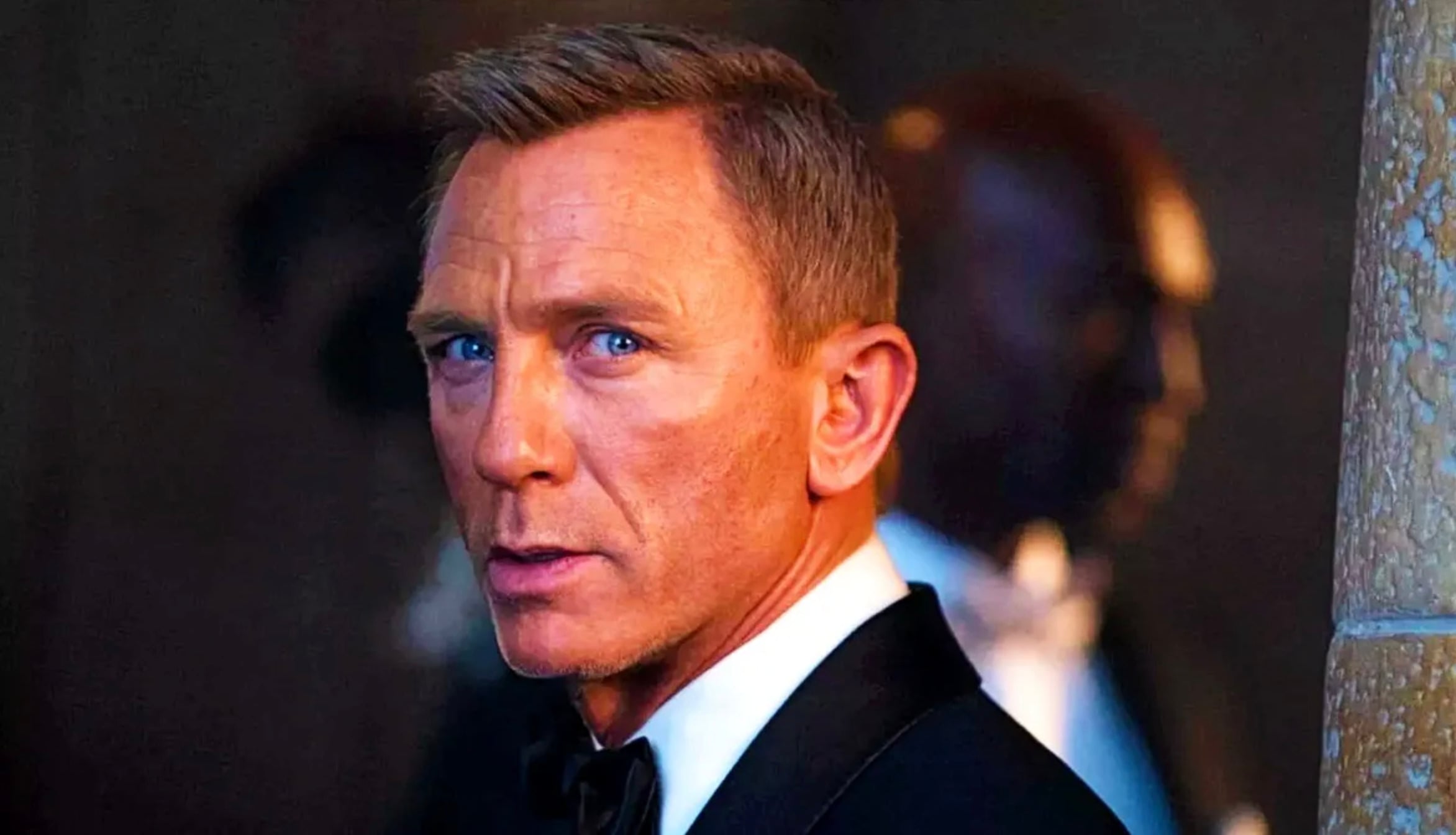 A man in a black tuxedo looks over his shoulder with a serious expression. The background is dimly lit, with another person in formal wear visible but out of focus.