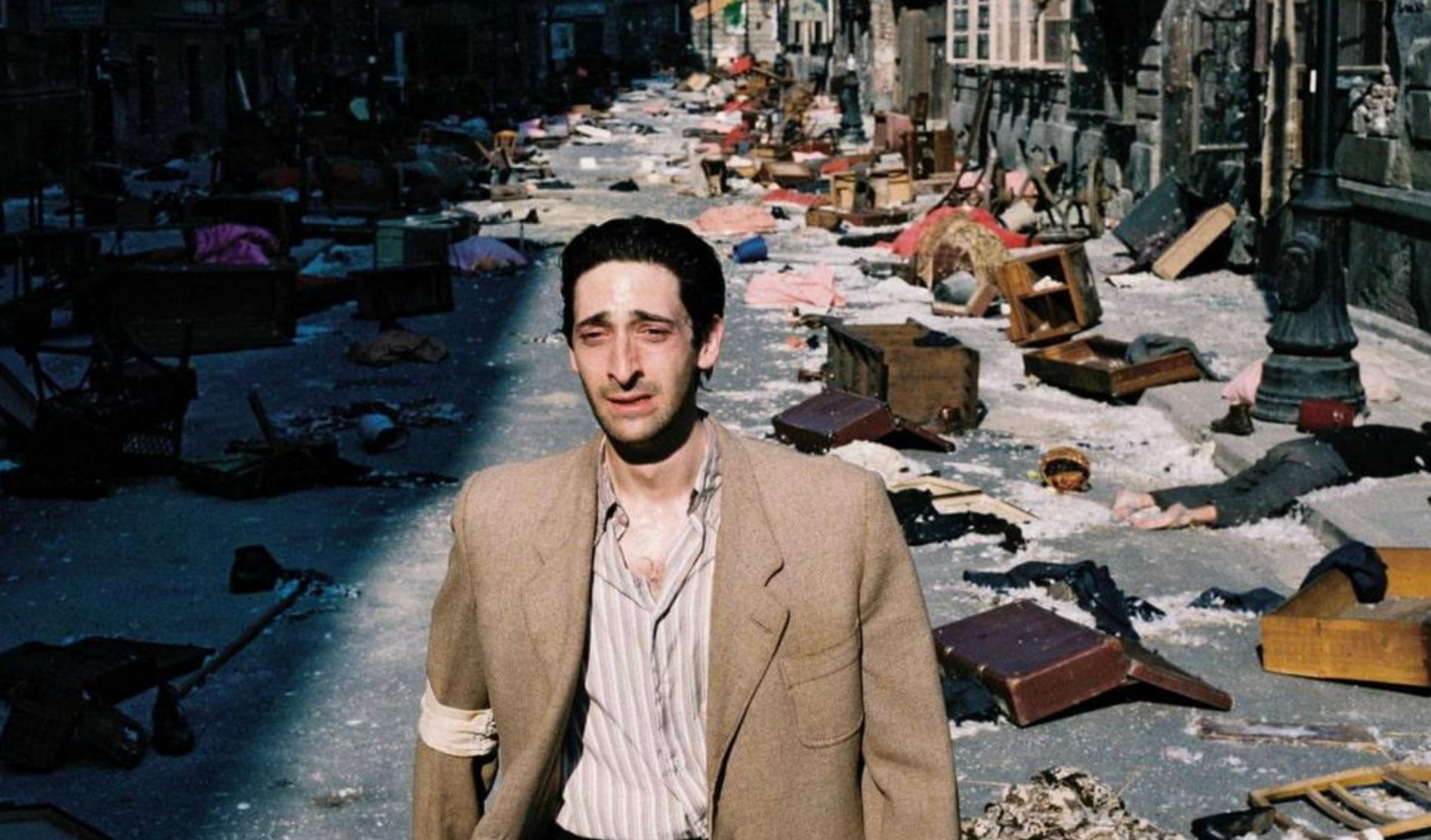 A man in a tattered suit stands in a sunlit street filled with scattered debris, overturned furniture, and belongings, with chaos and destruction visible in the background.