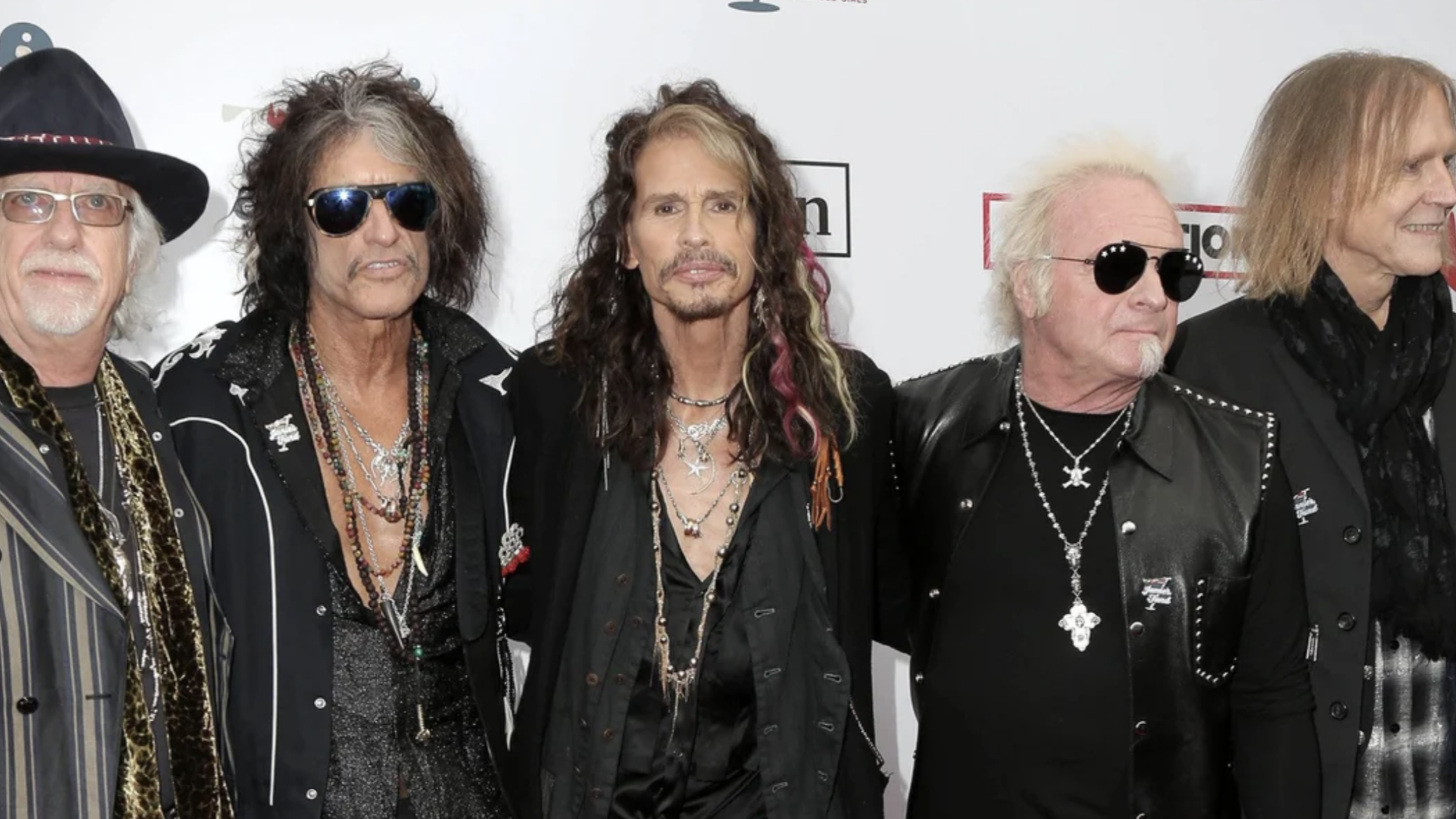 Five men stand together on a red carpet, wearing black clothing, sunglasses, and jewelry. They are posing for a group photo against a white backdrop with visible event logos.