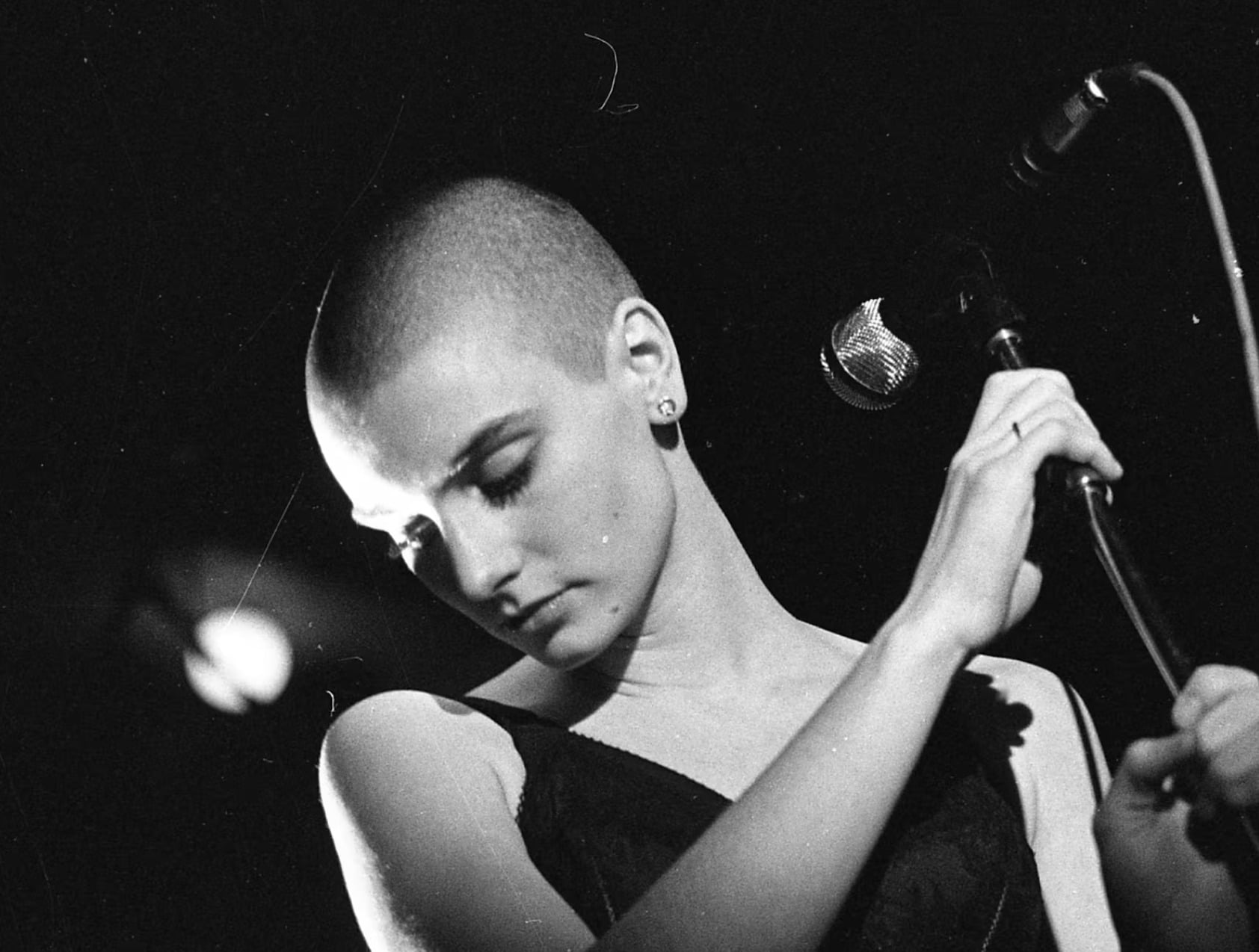 Black-and-white photo of a person with a shaved head, wearing a sleeveless top, standing in front of a microphone and looking downward with a calm, introspective expression.