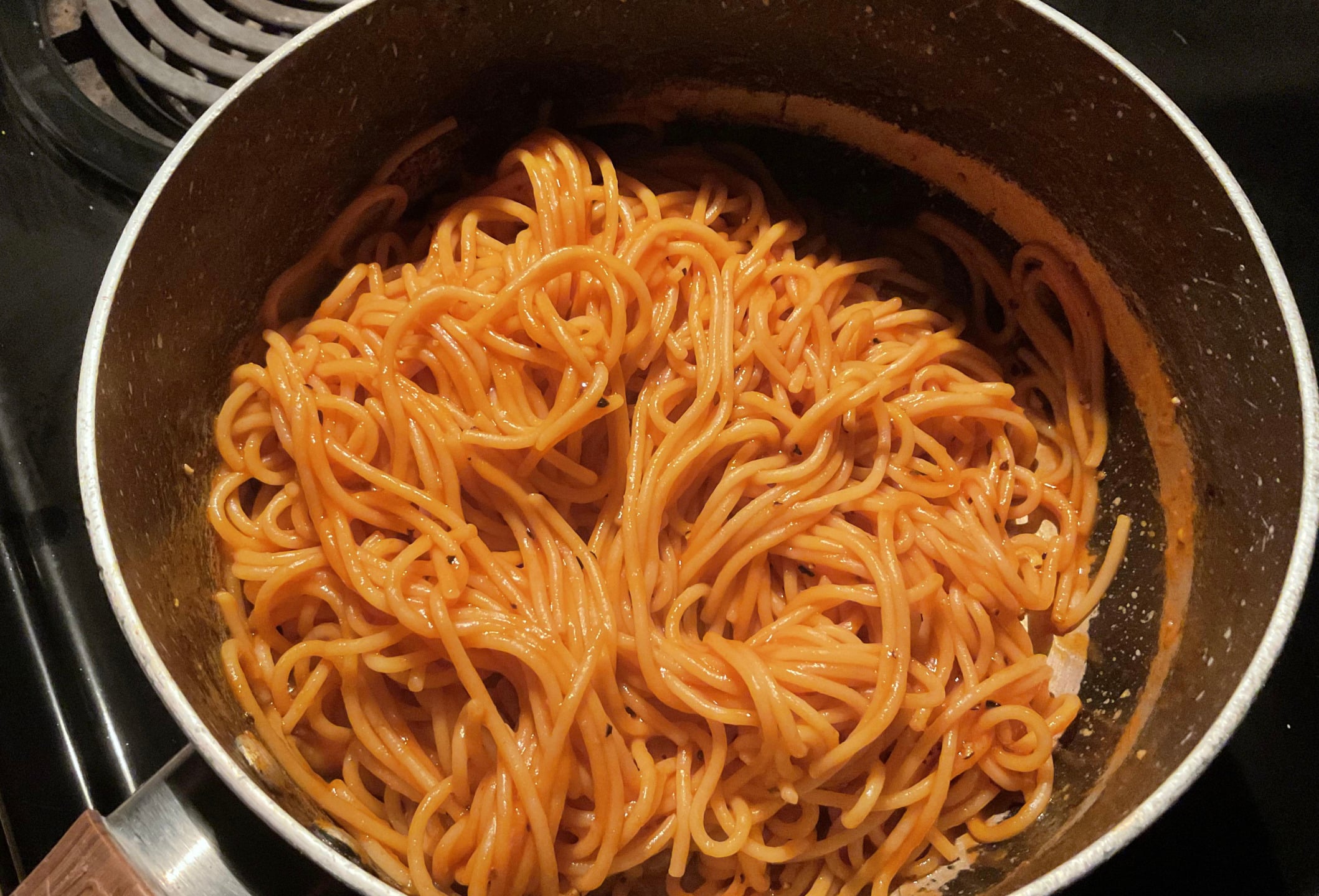 A pot filled with cooked spaghetti noodles coated in red tomato sauce, resting on a black stovetop.