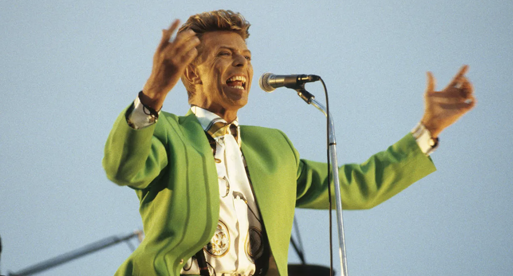 A singer wearing a bright green jacket performs energetically on stage, singing into a microphone and gesturing with both hands, with a clear blue sky in the background.