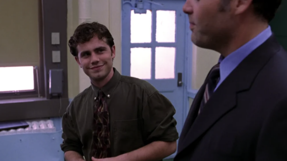 A young man in a green shirt and patterned tie stands smiling, looking at an older man in a suit and tie who is partially visible in the foreground; they appear to be conversing in a room with blue doors and windows.