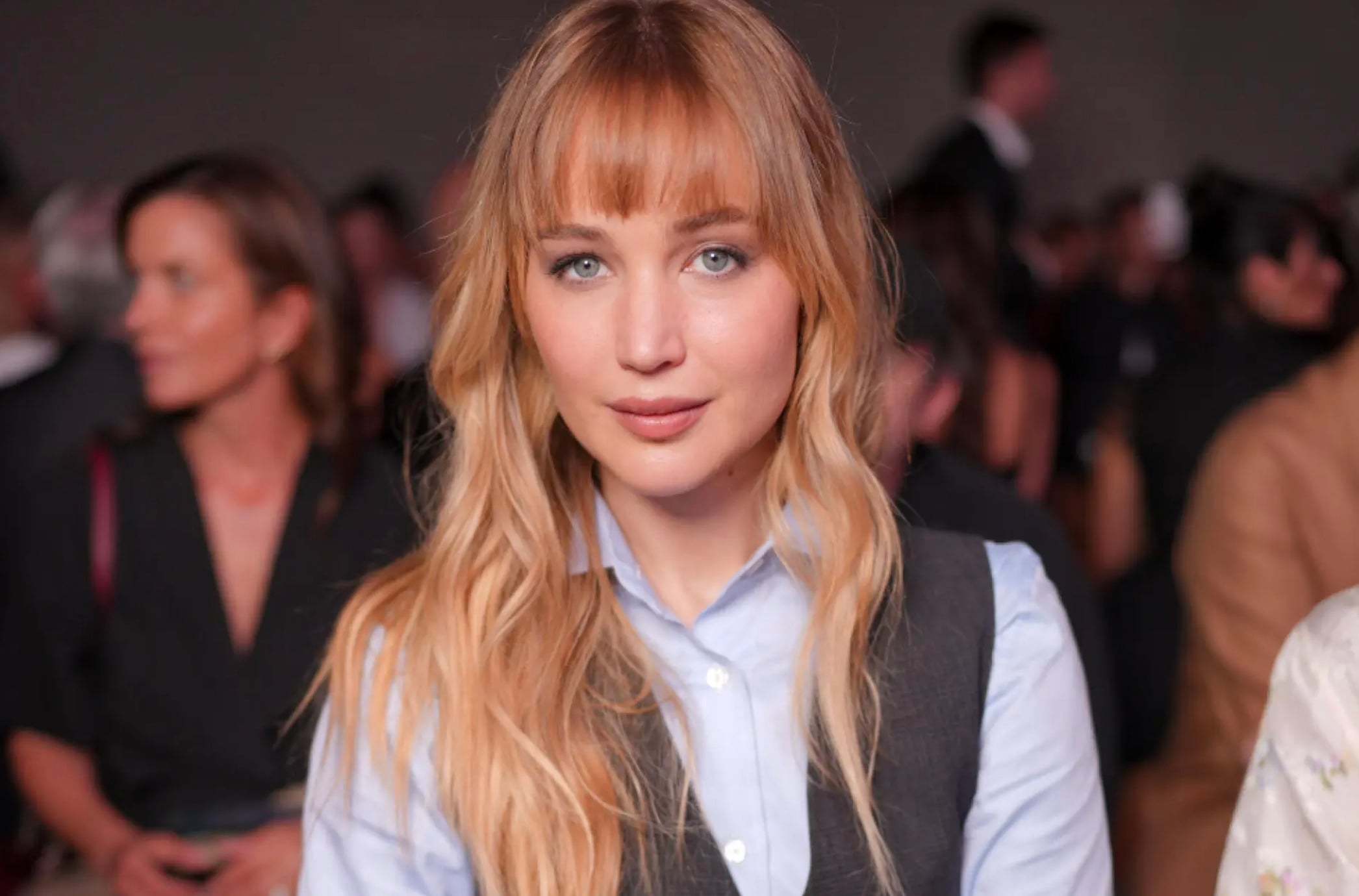 A woman with long blonde hair and bangs, wearing a light blue shirt and dark vest, sits indoors with a blurred crowd in the background. She is looking directly at the camera with a neutral expression.