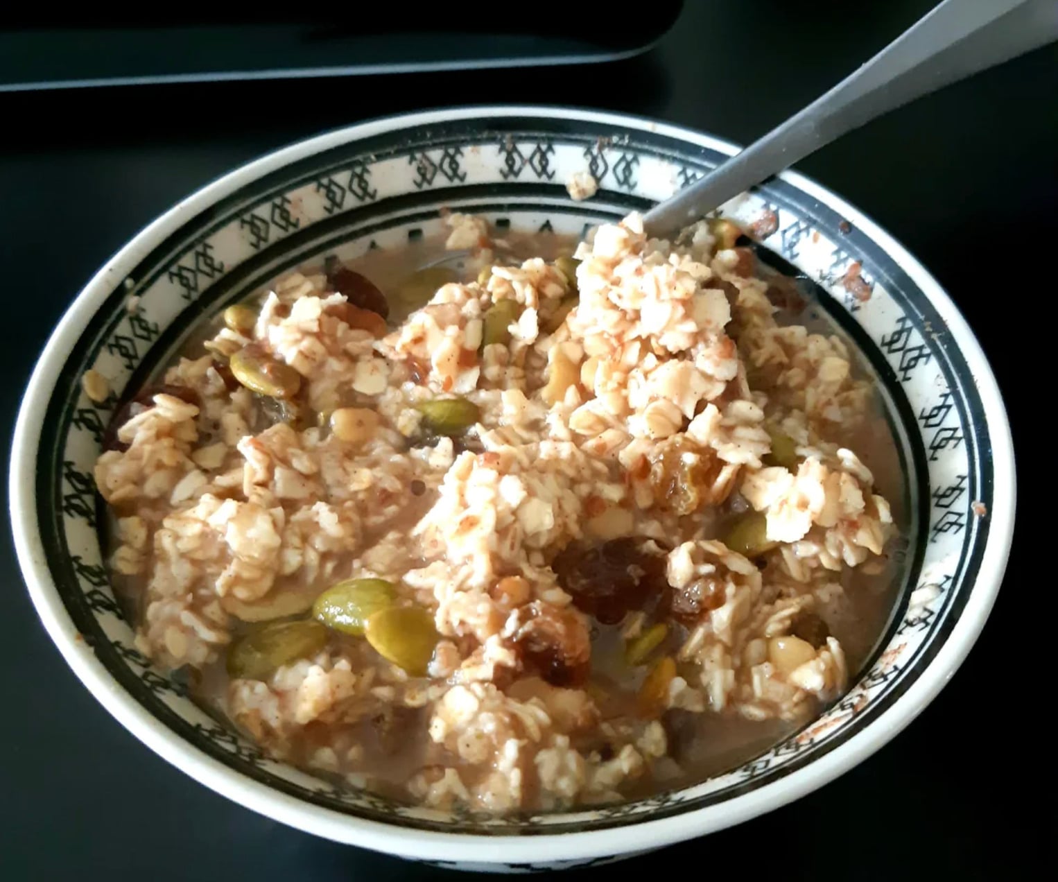 A patterned bowl filled with oatmeal mixed with raisins and pumpkin seeds, with a metal spoon resting inside, placed on a dark surface.