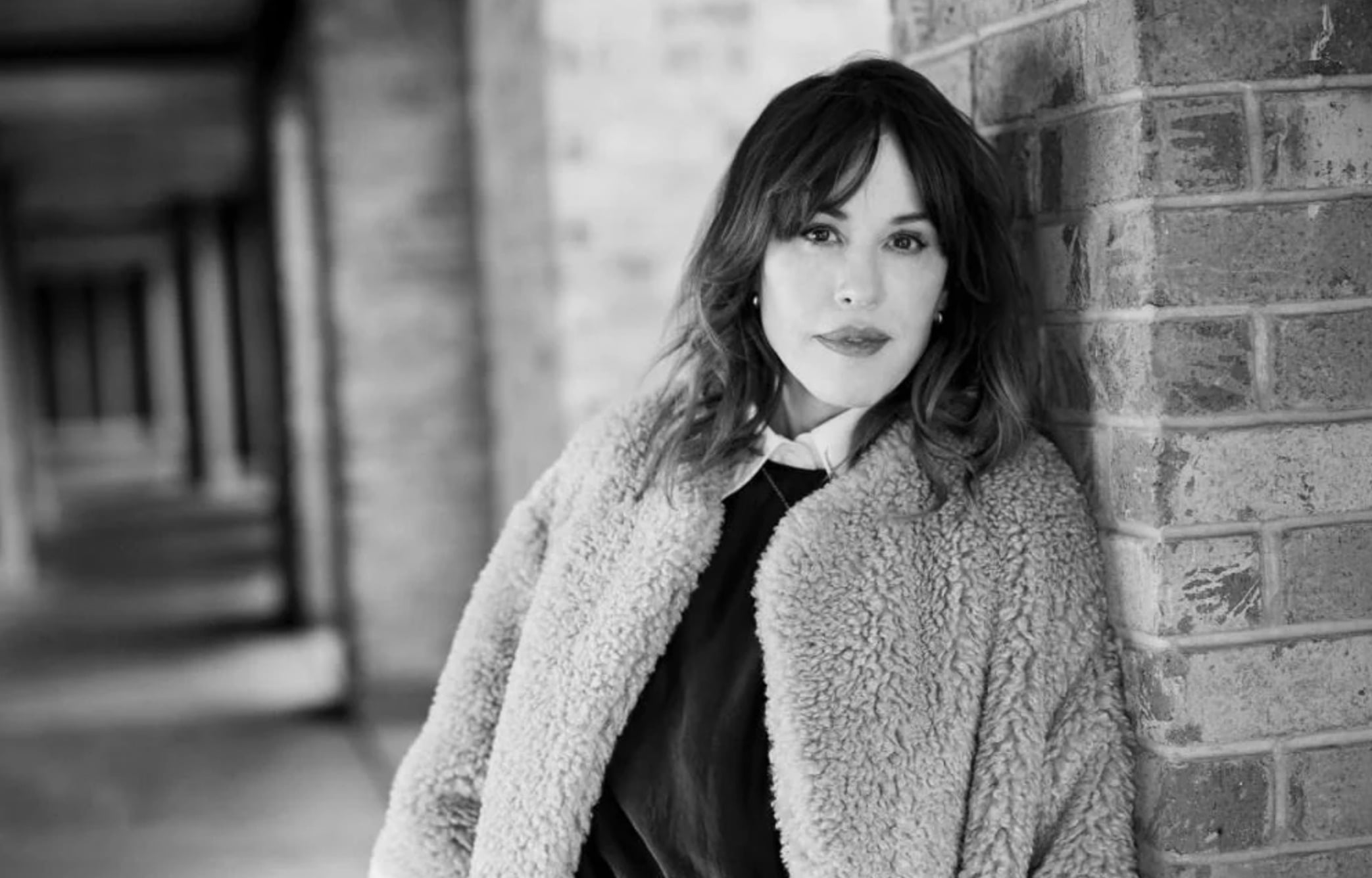 A woman with wavy hair wearing a textured, furry coat leans against a brick wall. The photo is in black and white, and the background shows a corridor with more brick walls.