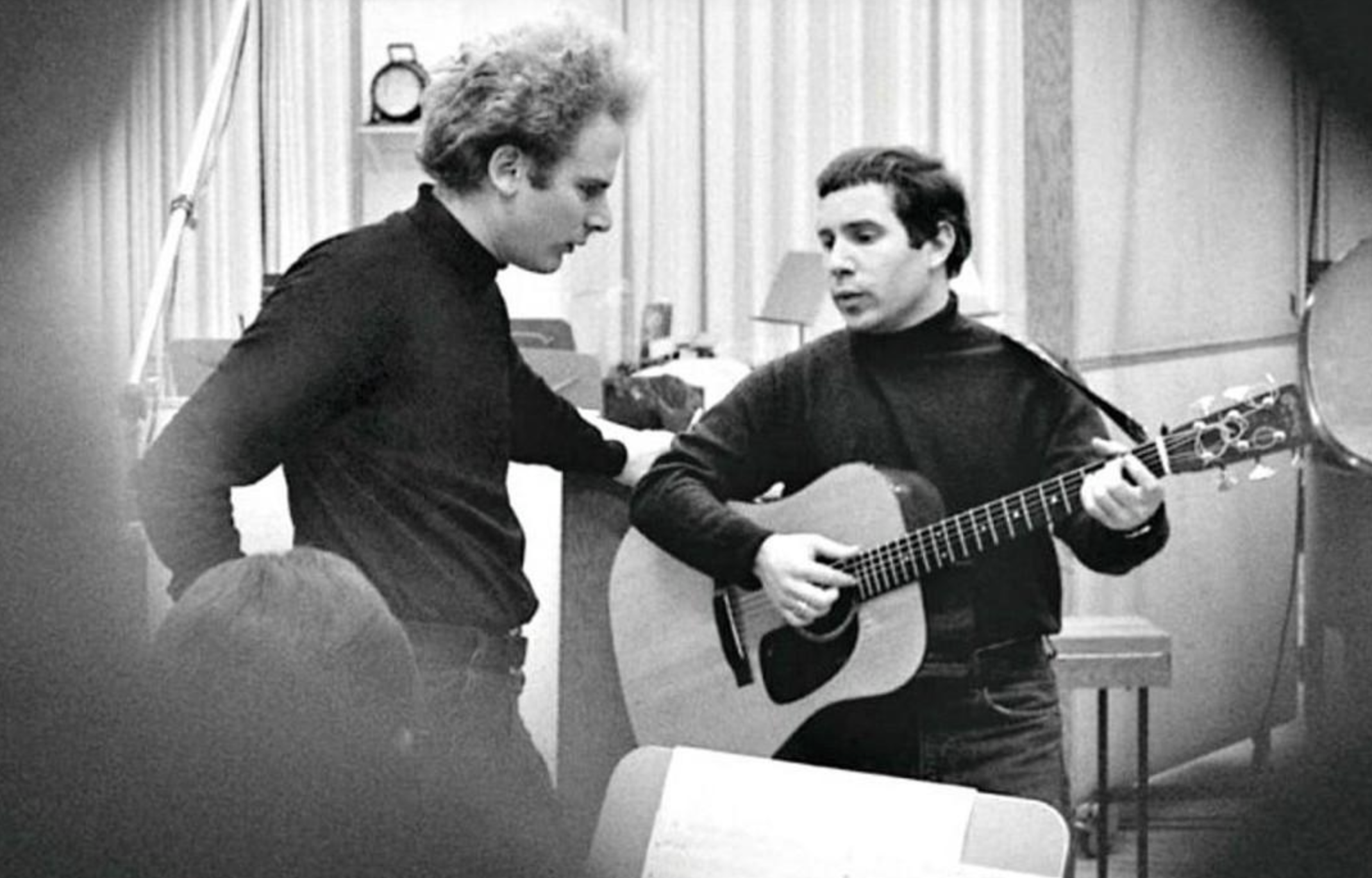 Two men in a recording studio, one standing and singing, the other playing an acoustic guitar. Both wear dark turtlenecks and appear focused, with musical equipment and sheets visible in the background.