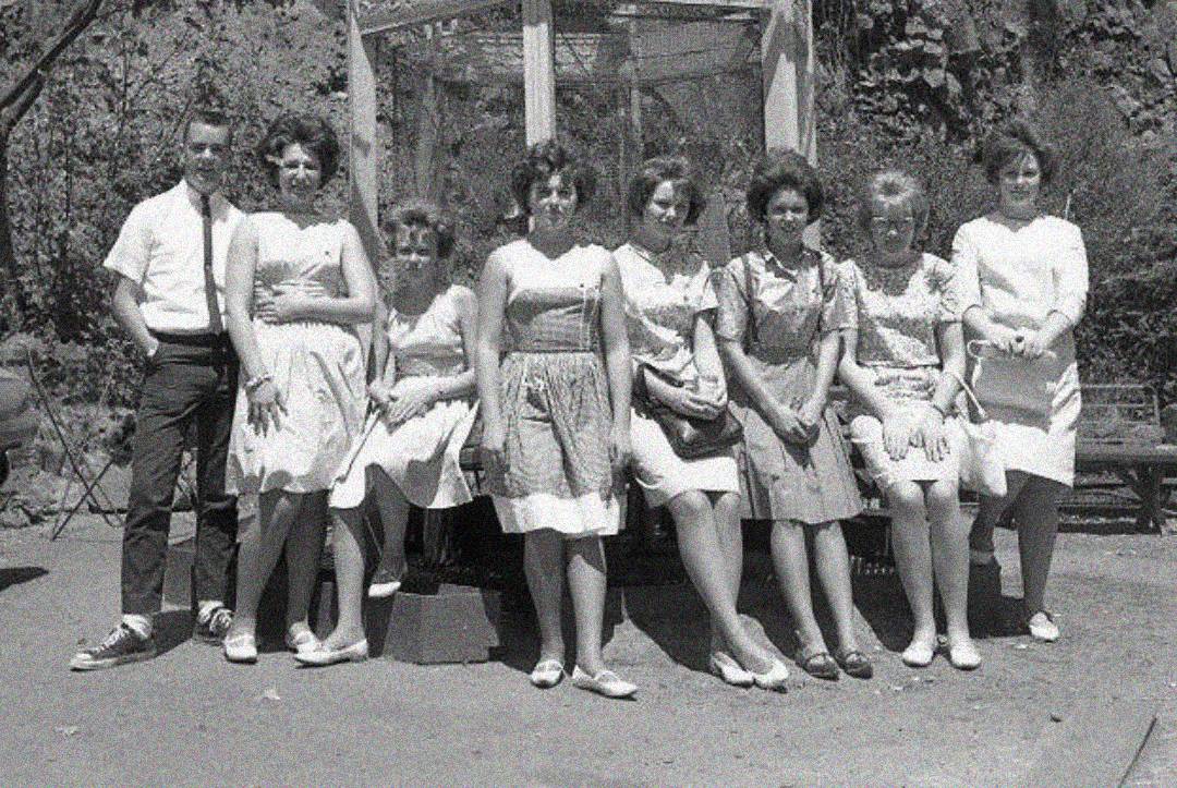 A vintage black and white photo of eight young adults, one man and seven women, standing and sitting outdoors in front of a structure, all dressed in light summer clothes and smiling at the camera.