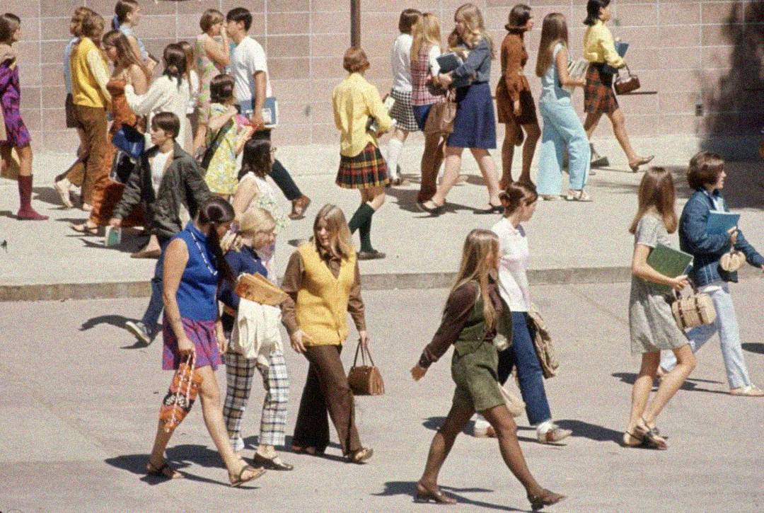 A group of students in 1970s-style clothing walk in different directions outside a school building, carrying books and bags. The clothing includes bright colors, patterns, and knee-high socks.