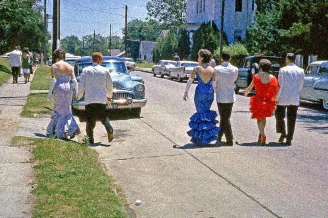 A group of young people dressed in formal attire, including colorful dresses and suits, walk down a suburban sidewalk beside vintage cars on a sunny day.