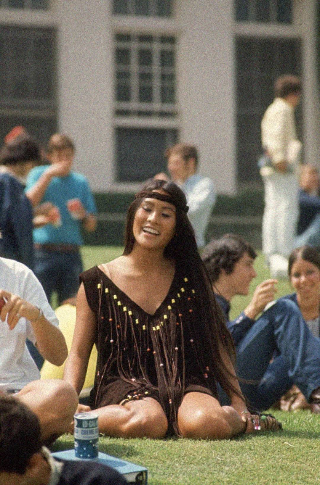 A young woman in a brown fringed dress with colorful beads sits cross-legged on grass, smiling, at an outdoor gathering with other casually dressed people in the background.