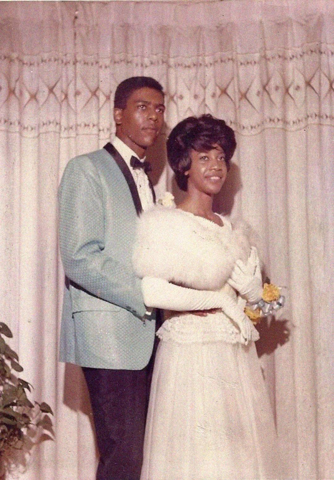 A young couple dressed formally stands together; the man wears a light blue tuxedo jacket with a black bow tie, and the woman wears a white gown with gloves, holding a fur stole. They pose in front of a patterned curtain.