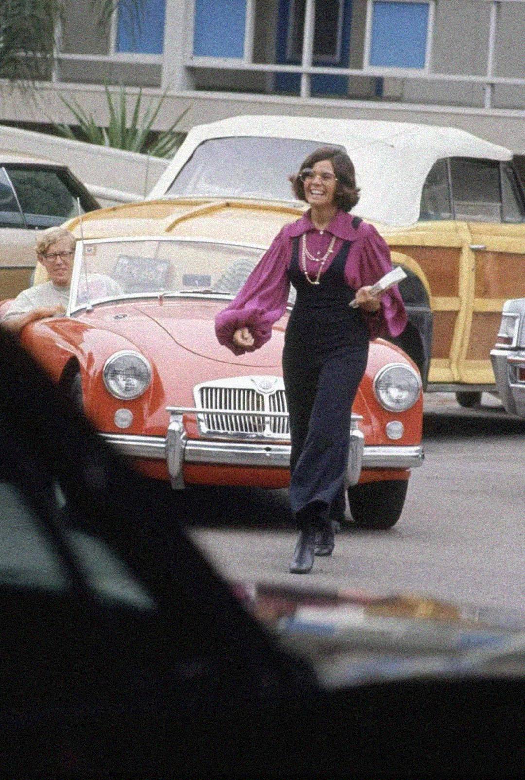 A woman in a magenta blouse and dark overalls walks in a parking lot, smiling and holding papers, near a classic red convertible with a man sitting inside. Vintage cars are parked in the background.