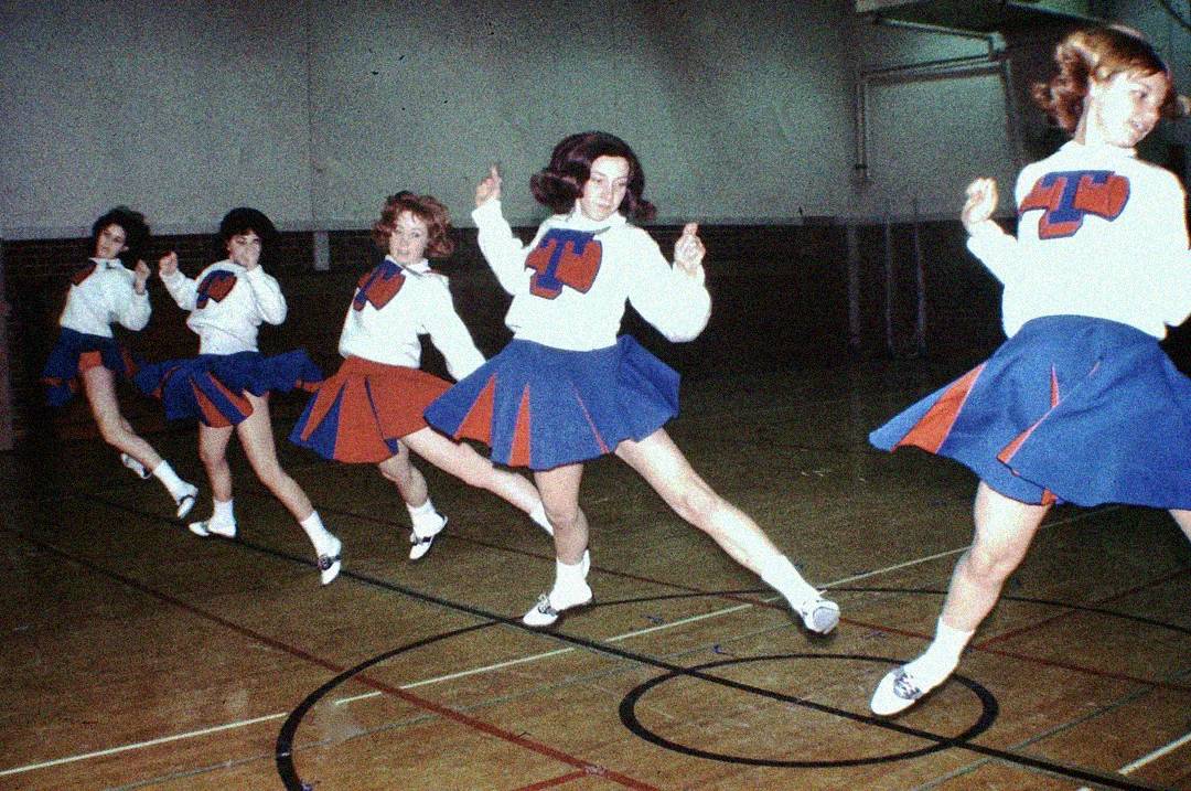 Five cheerleaders in matching blue and orange uniforms perform a synchronized high-kick routine in a gymnasium with a wooden floor and brick walls.