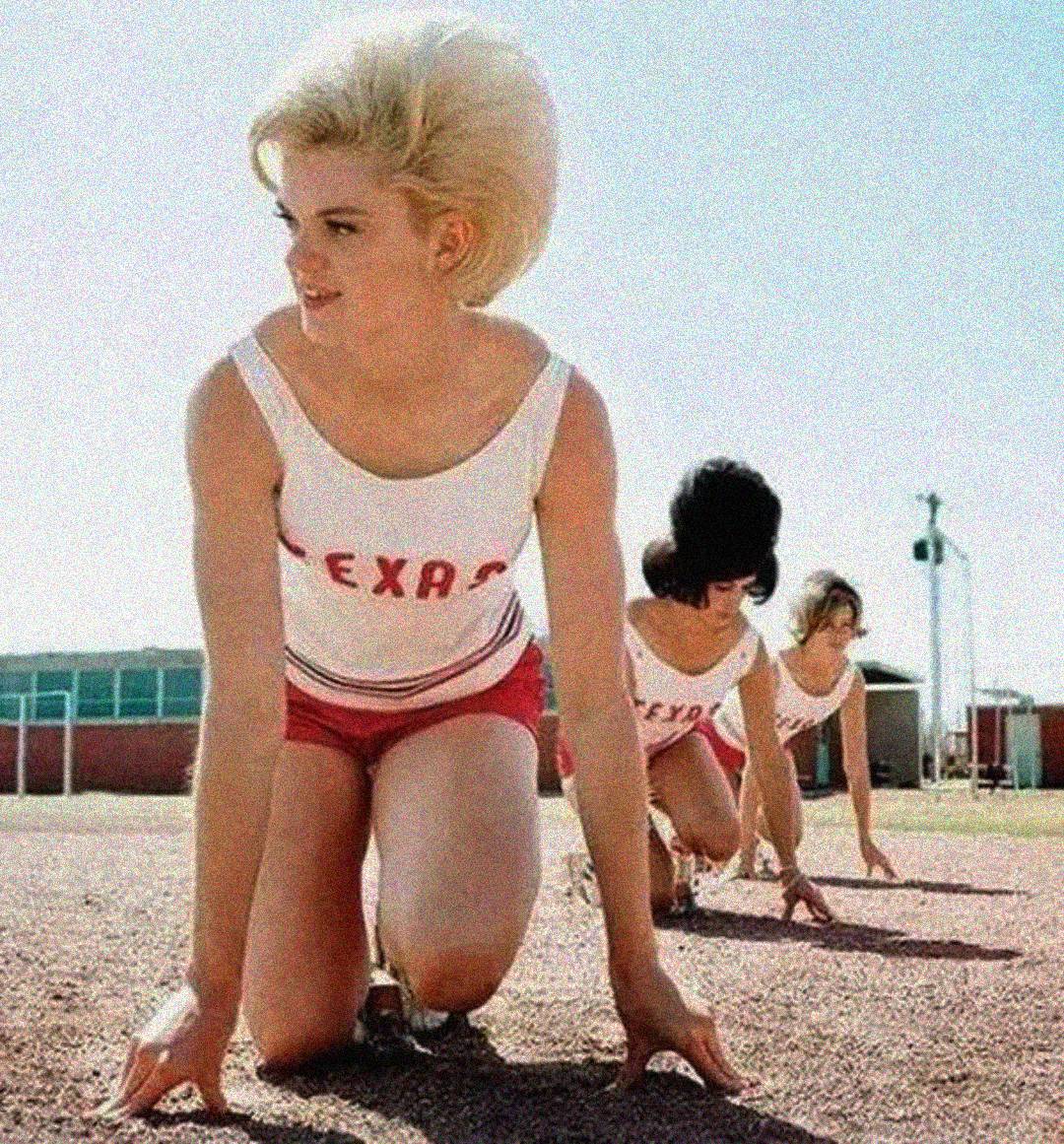 Three women in athletic uniforms that say "TEXAS" are crouched at a starting line outdoors on a sunny day, ready to race on a track. The woman closest to the camera has short blond hair.