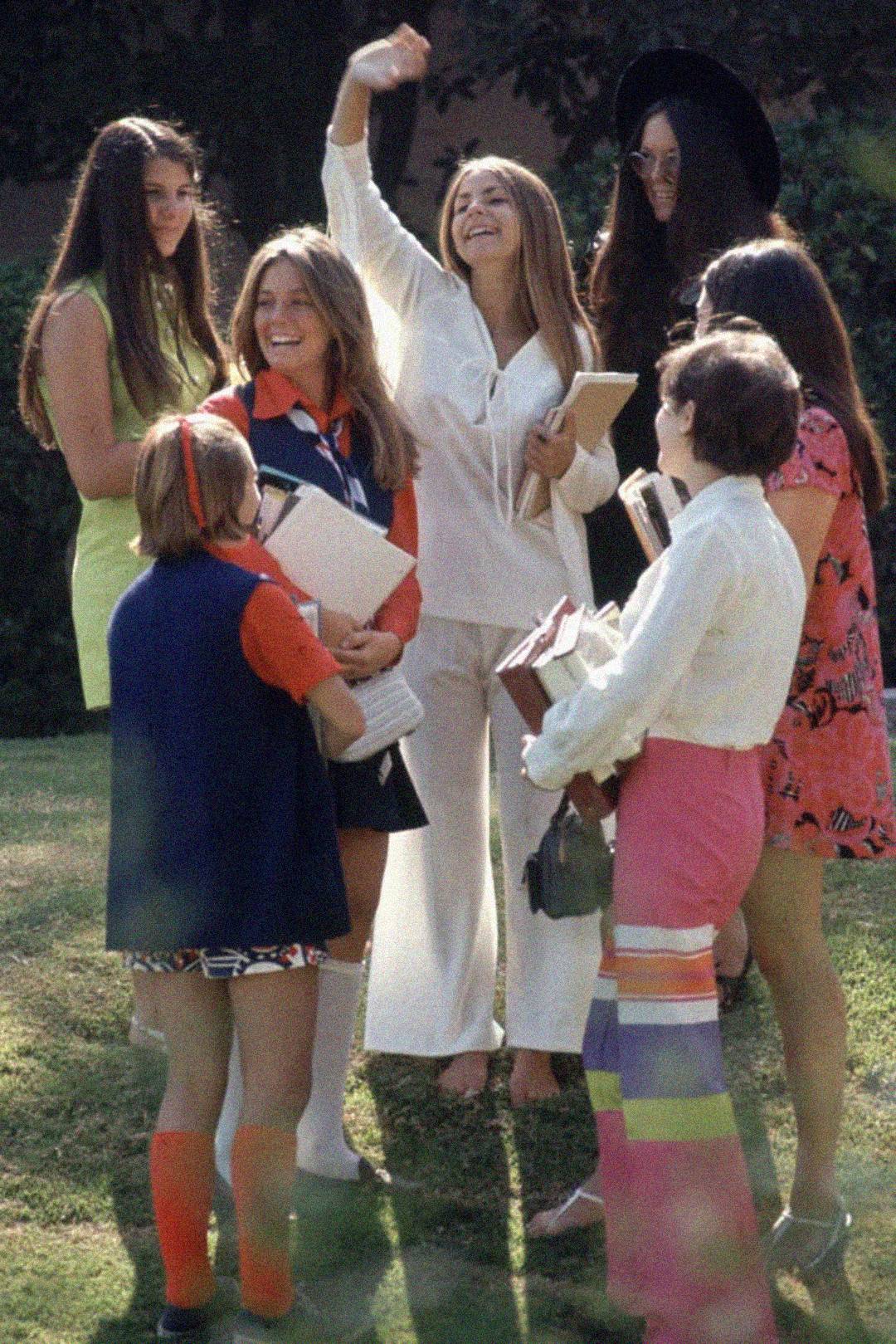 A group of young women in colorful 1970s clothing stand together outdoors, smiling and talking while holding books. One woman in white raises her arm and waves in a cheerful gesture. Trees and grass are visible in the background.