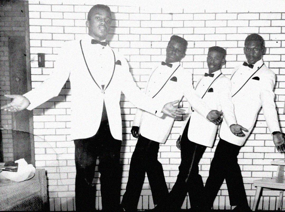 Four men wearing matching white tuxedo jackets and bow ties pose in front of a brick wall. Three lean to the right in unison, while one stands in front with arms extended, all appearing to perform or dance.