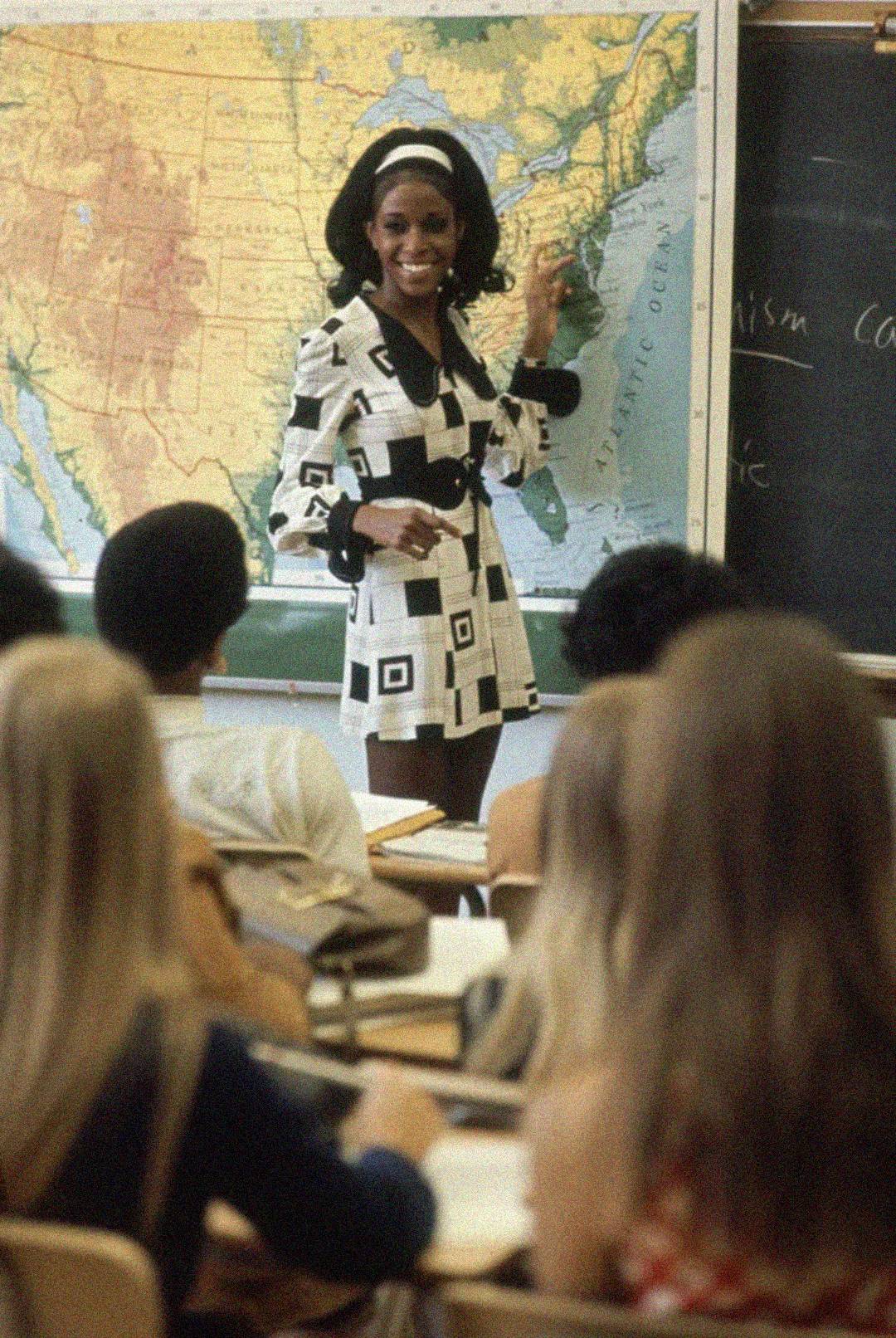 A woman in a black and white patterned dress stands in front of a U.S. map and chalkboard, smiling and pointing, while students seated at desks watch her in a classroom.