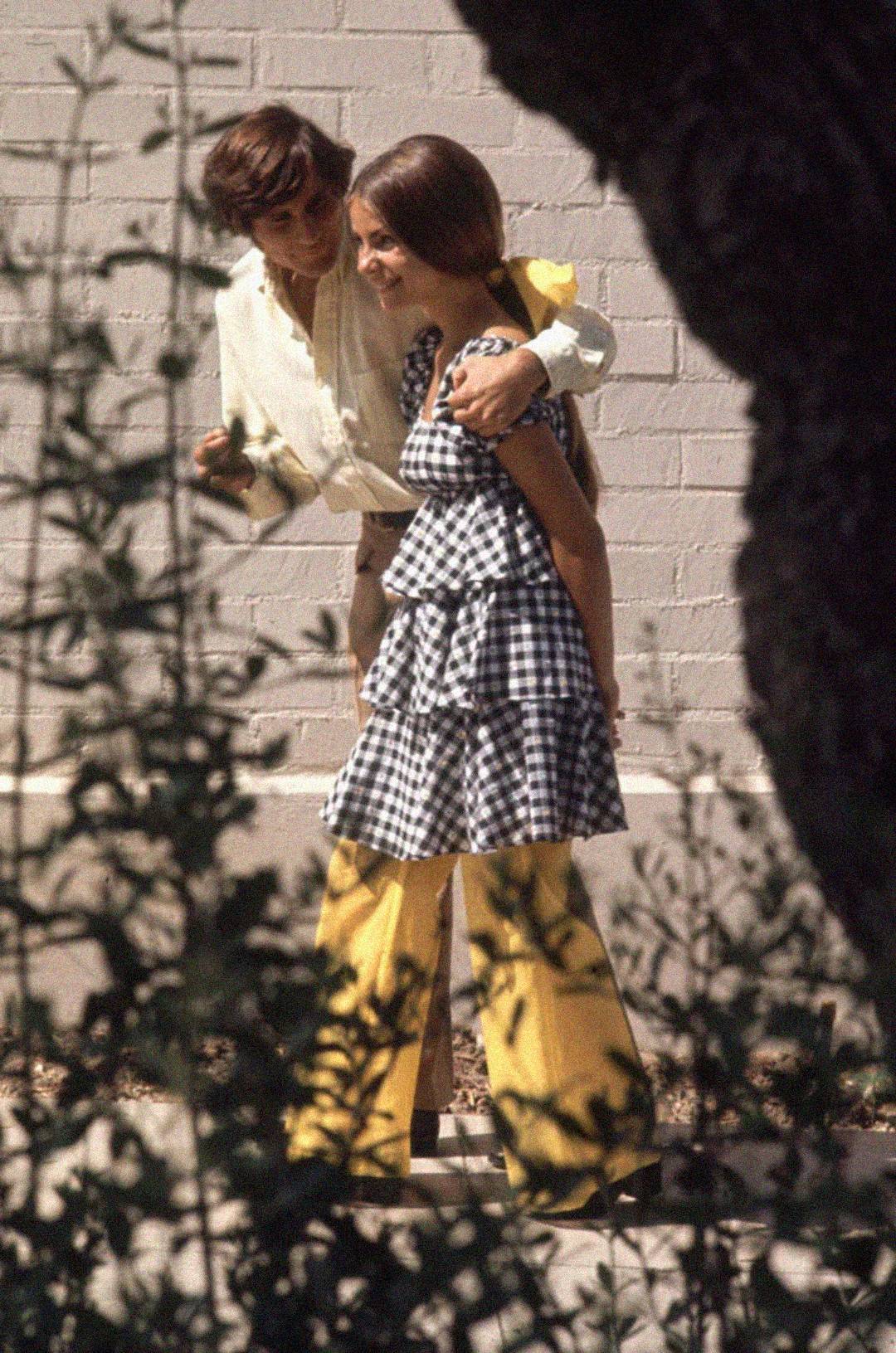 A young man with an arm around a smiling young woman walk outdoors. The man wears a light shirt and yellow pants, while the woman wears a black-and-white checkered dress over yellow pants. Trees and branches frame the image.