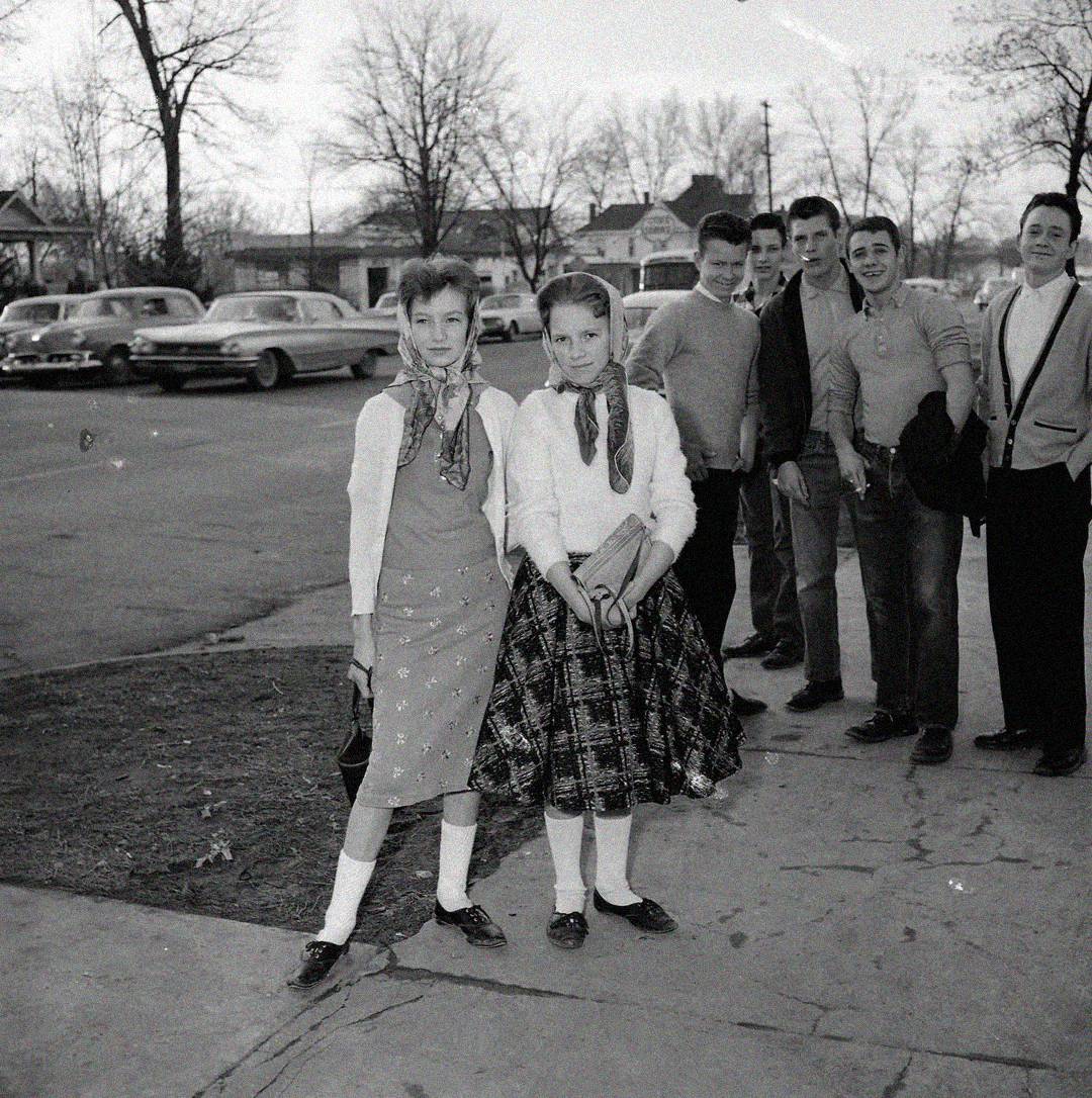 Two girls wearing scarves, sweaters, skirts, and saddle shoes stand in the foreground, while a group of young men in 1950s-style clothing stand behind them on a street lined with parked vintage cars and bare trees.