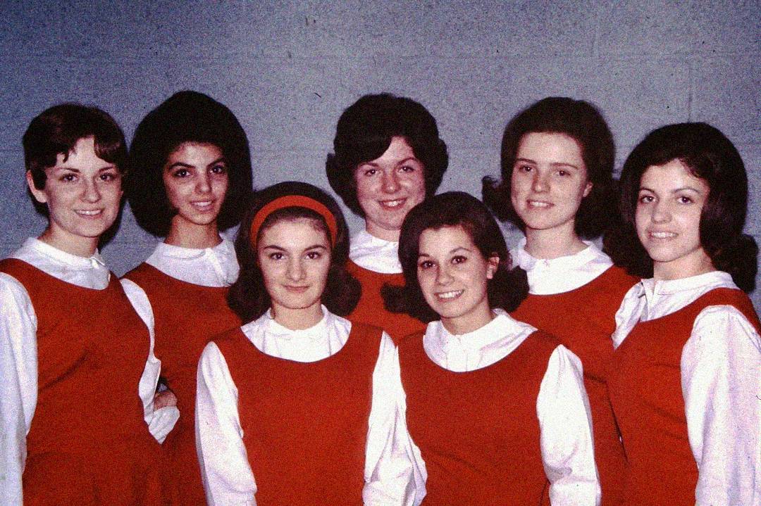 Seven young women stand closely together, smiling at the camera. They are all wearing matching red jumpers over white blouses, and have neatly styled dark hair. The background is a plain, light-colored wall.