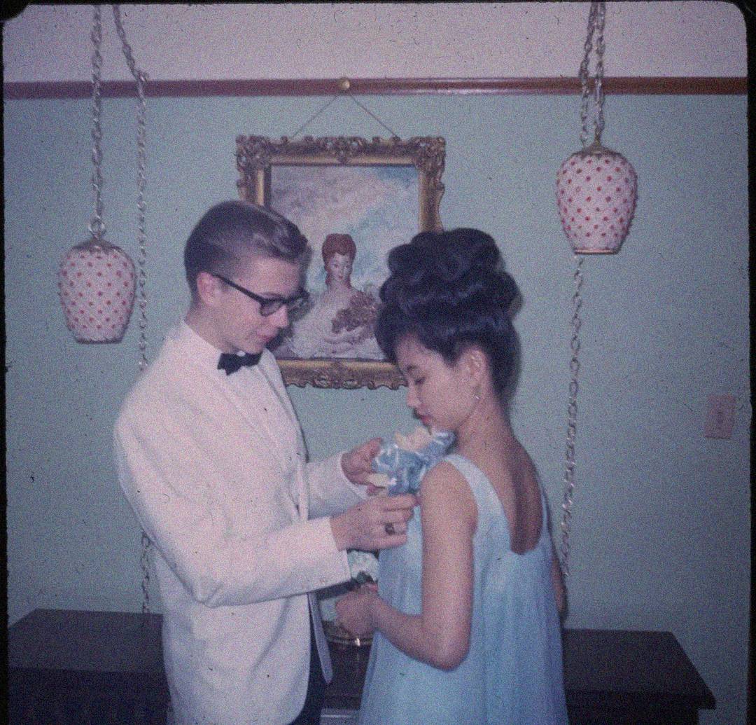 A young man in a white tuxedo pins a corsage on a young woman in a blue dress. They stand in front of a wall with hanging lamps and a framed painting of a woman in the background.
