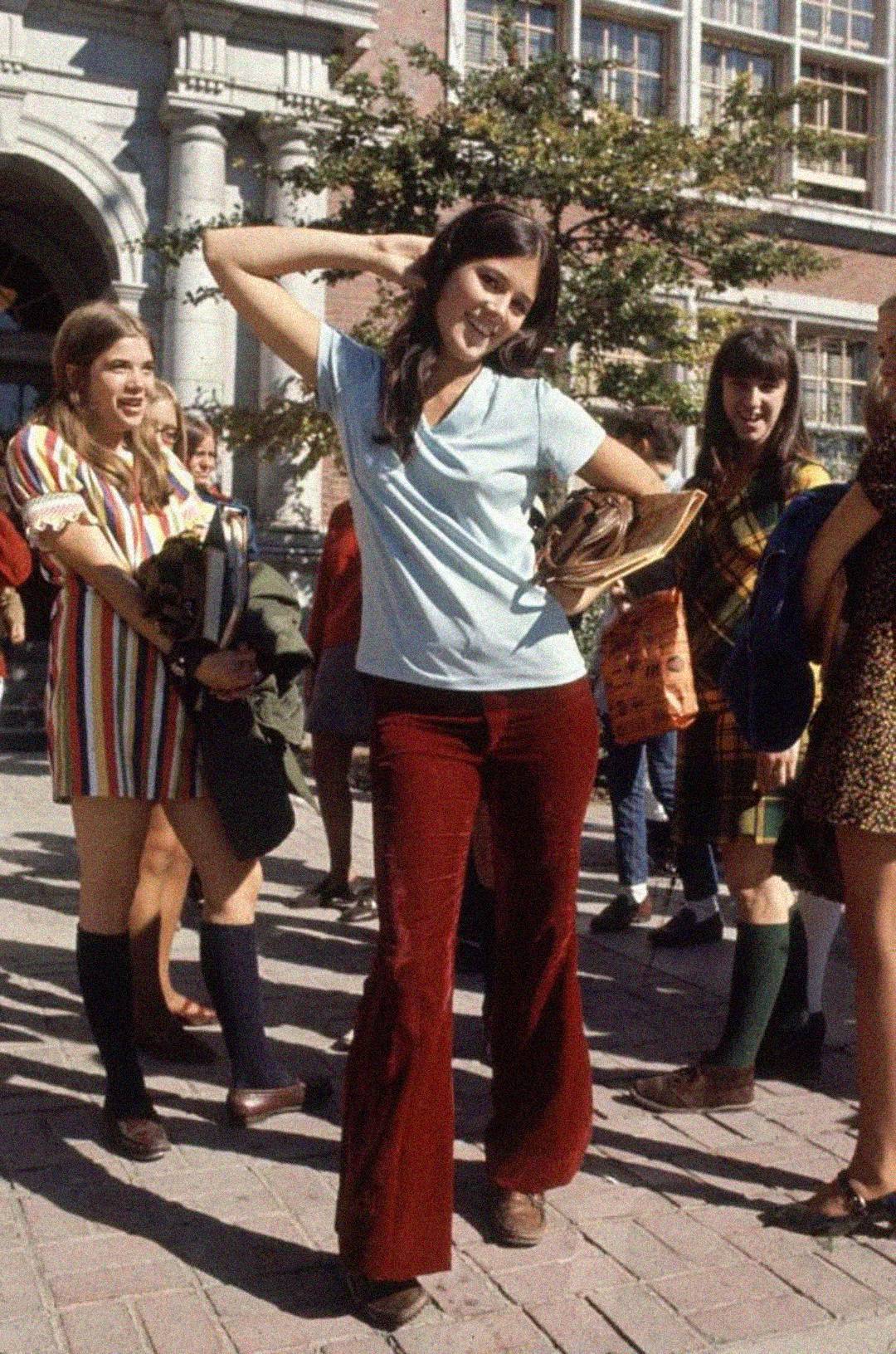 A young woman in a light blue shirt and red bell-bottom pants poses playfully outside a school, surrounded by other smiling students in colorful, retro 1970s clothing.
