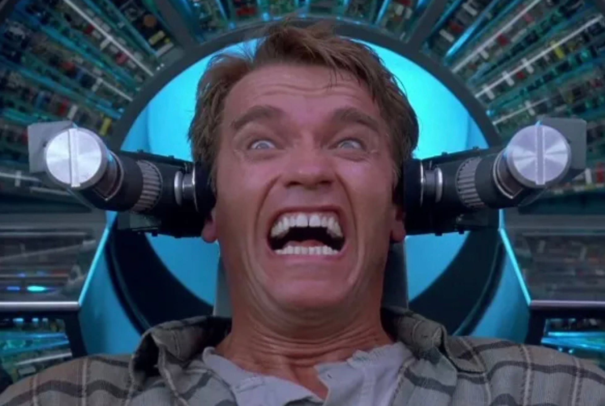 A man with a terrified expression sits in a futuristic machine with metal devices aimed at his head, surrounded by intricate electronic components and glowing blue light.
