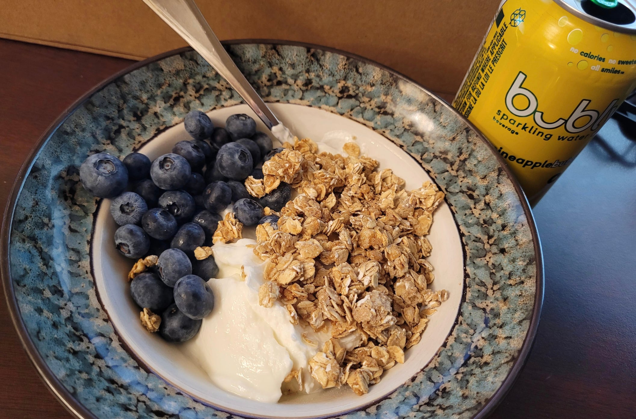 A bowl with yogurt, granola, and fresh blueberries, with a spoon resting inside. Next to the bowl is a can of Bubly pineapple sparkling water on a wooden surface.