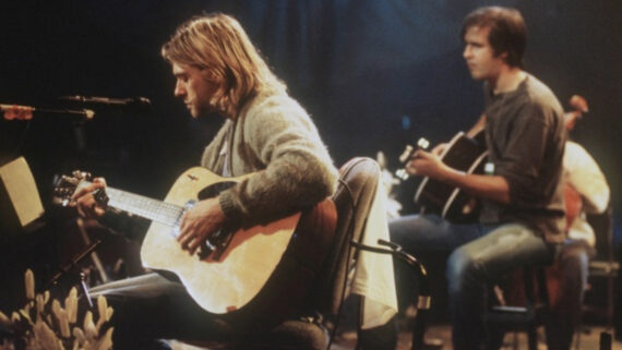 Two musicians sit on stage playing acoustic guitars under soft stage lighting. One wears a gray cardigan and has long blond hair, the other wears a t-shirt and jeans. Music stands and equipment are visible around them.