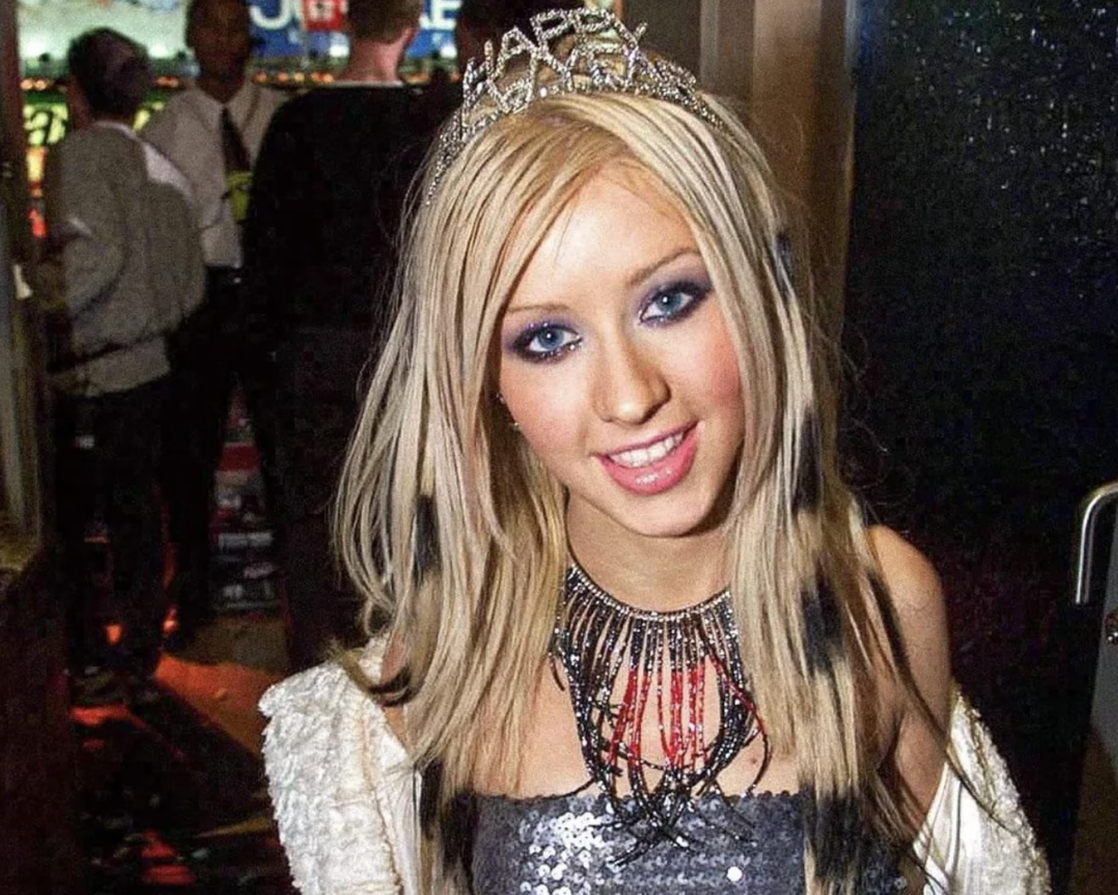 A woman with long blonde hair wearing a tiara, heavy blue eye makeup, a sparkly silver top, a bold necklace, and a white jacket stands indoors, smiling at the camera with people blurred in the background.
