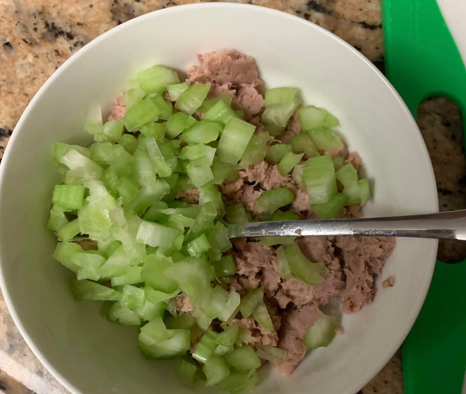 A bowl containing chopped celery and flaked tuna with a spoon resting inside, placed on a granite countertop.