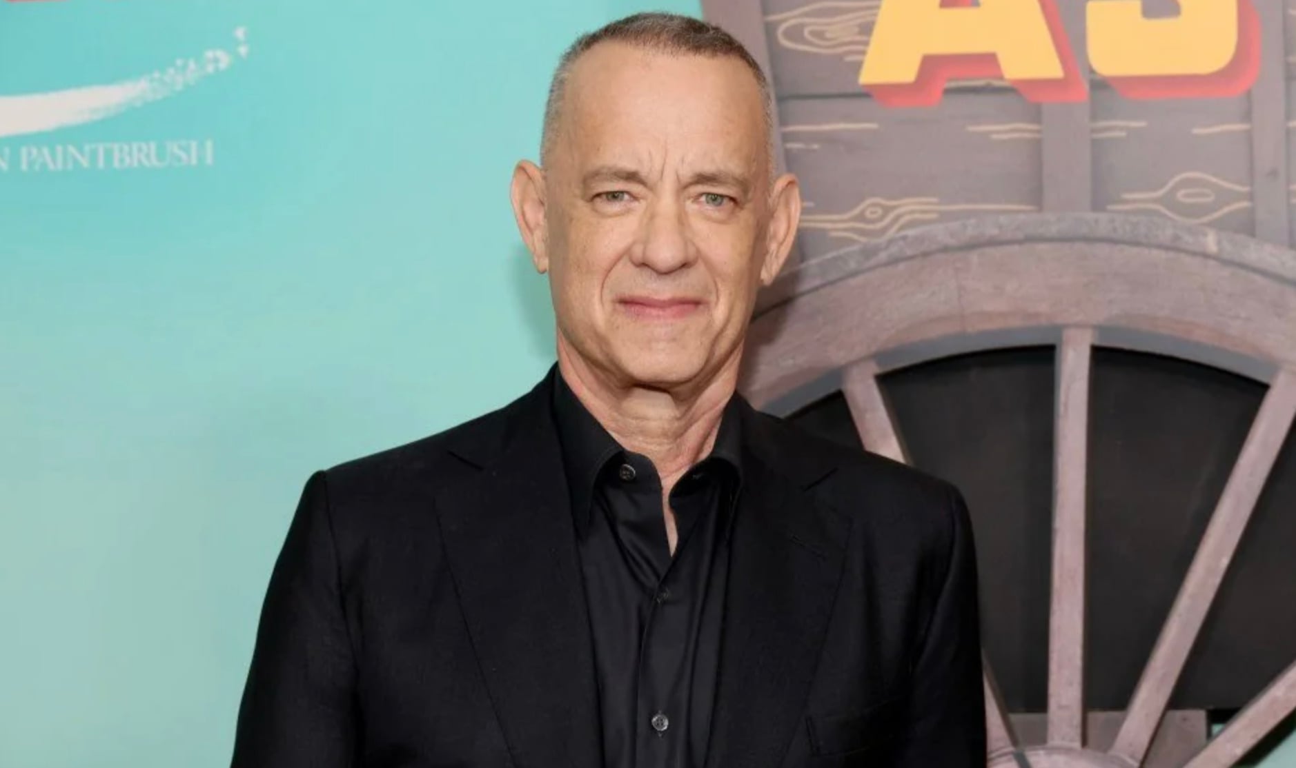 A man with short gray hair, wearing a black suit and black shirt, stands in front of a colorful backdrop featuring part of a wagon wheel and bold yellow lettering.