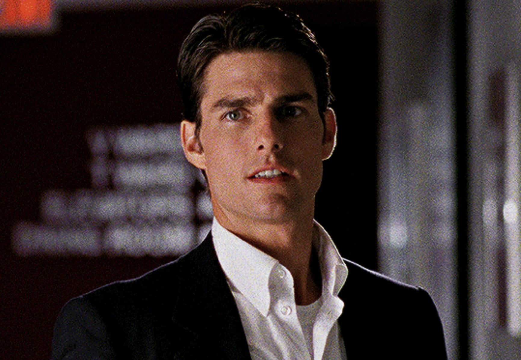 A man with short dark hair wearing a white dress shirt and black blazer stands indoors, looking slightly to the side with a serious expression. The background is blurred and includes some text on a wall.
