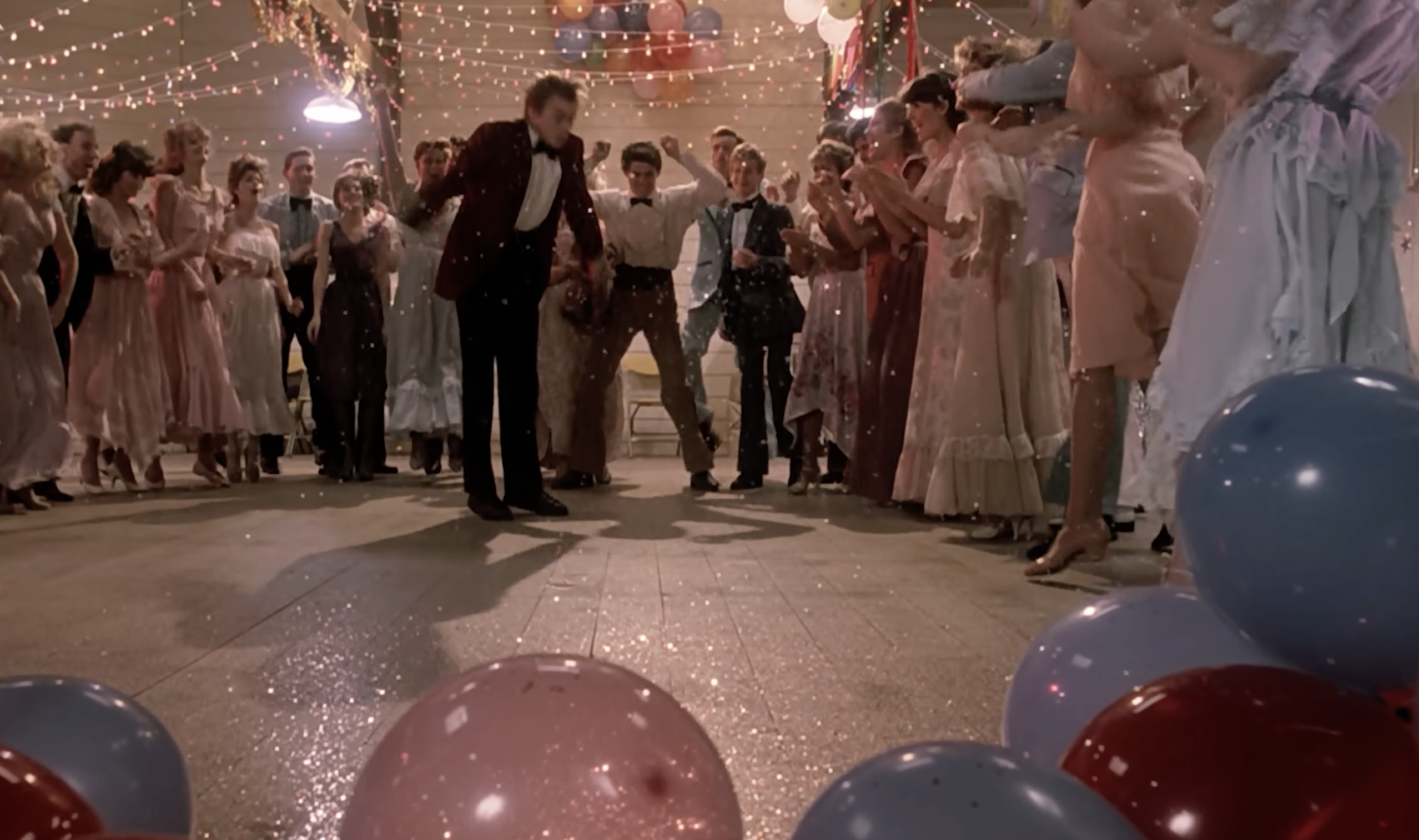 A group of people in formal attire stand in a decorated hall with balloons and string lights, cheering as a man in a maroon jacket bends forward near the dance floor. Balloons and confetti are scattered on the floor.